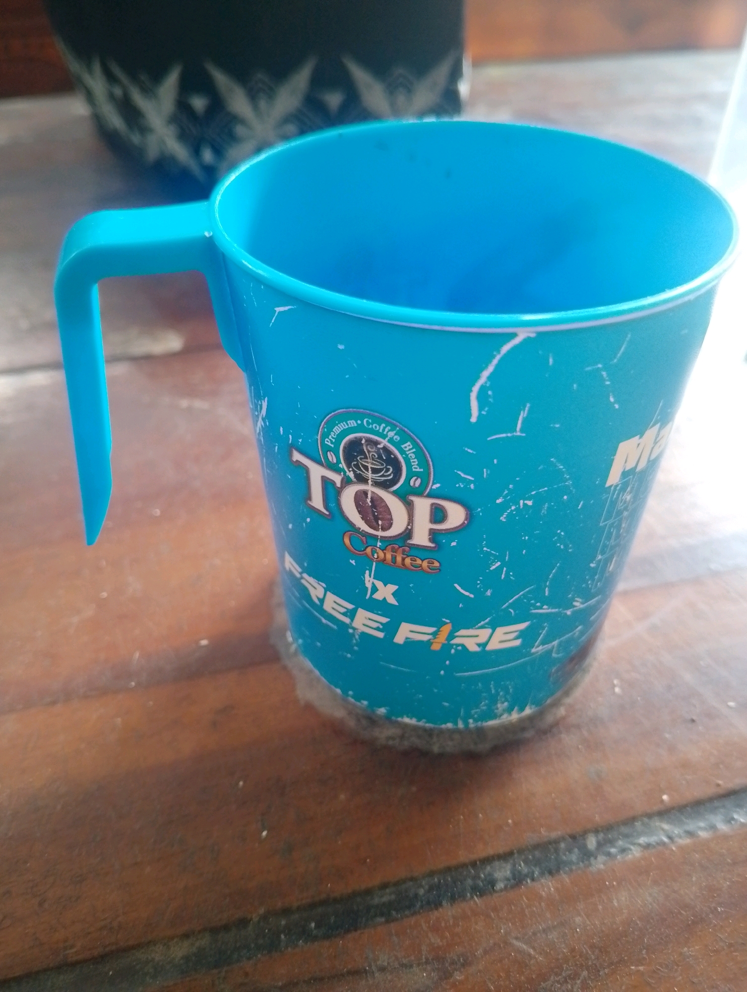 A blue plastic mug with the "Top Coffee" logo and "Free Fire" branding is situated on a wooden table. The mug has a handle and appears to be slightly worn with some scratches and marks. In the background, a portion of a dark, patterned object is visible. The table surface is made of wooden planks with visible grain and some scattered dust. The image was captured in Kendalrejo, Indonesia.