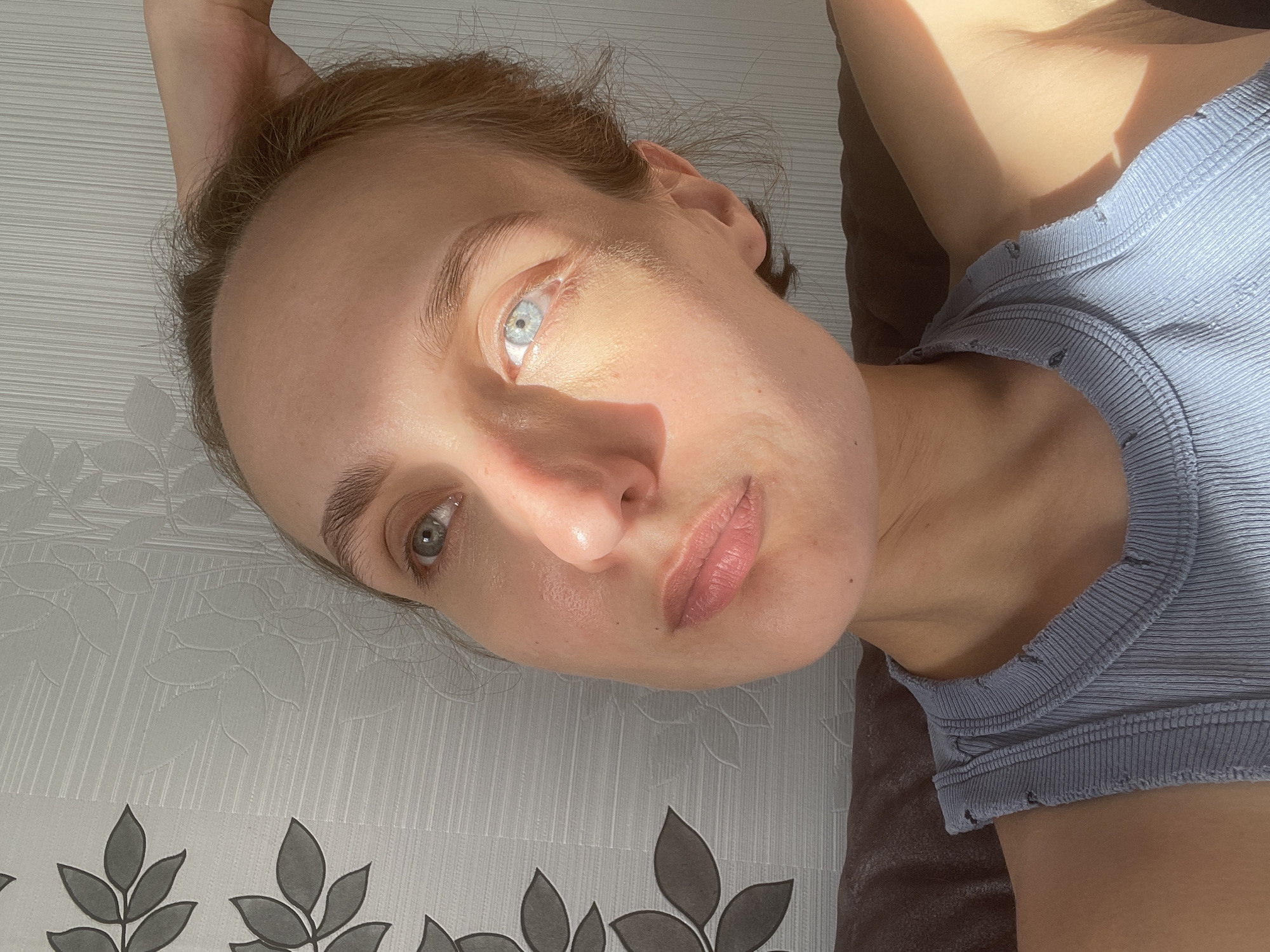 The image presents a close-up, high-angle portrait of a fair-skinned individual with light brown hair and light blue/grey eyes, lying down and looking upwards towards the camera. The person is wearing a light blue, ribbed tank top or camisole, featuring visible stitching and small, distressed details along its edges.
Strong, natural sunlight streams into the scene, creating dramatic highlights and shadows across the face and upper body. One eye is brightly illuminated, while other areas, such as the chin and part of the forehead, are in shadow, suggesting a specific time of day, likely morning or late afternoon. The individual's expression is neutral and calm.
The background directly beneath the head consists of a light-colored, subtly textured surface, possibly a bedsheet or wallpaper, with a repeating pattern of stylized dark grey leaf shapes along the bottom edge. To the right of the frame, underneath the person's upper body, a darker, textured surface, perhaps a brown pillow or blanket, is visible. There is no visible text. The setting is an indoor environment in Sharkan, Russia.