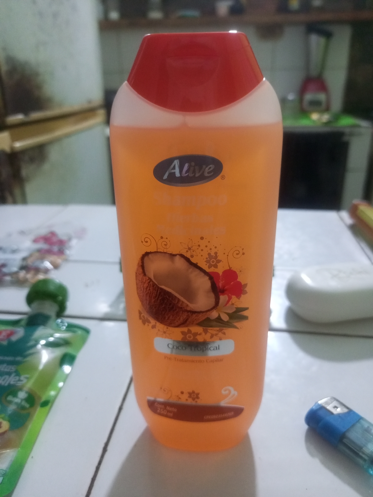 The image captures a close-up, eye-level shot of a bright orange shampoo bottle with a red cap, centrally positioned on a white tiled surface. The bottle features a prominent oval label with the brand name "Alive" in white text and a graphic of a halved coconut with tropical flowers. Text below the coconut reads "Coco Tropical" and "Pre-Tratamiento Capilar". Towards the bottom of the bottle, a rectangular label indicates "Cont. Neto 250ml".
To the left of the shampoo bottle, a portion of a green pouch with colorful graphics and text that appears to read "Frutas" and "Aples" is visible. To the right of the shampoo, a white bar of soap is partially in view, and further right, a blue and clear plastic lighter rests on the tiled surface.
The background is blurred but suggests an indoor setting. A light-colored refrigerator or similar appliance is visible on the left, and shelves with various objects, possibly kitchen items or appliances like a blender, are present in the upper background. The overall lighting is somewhat dim, with light sources likely originating from above and slightly to the right. The scene appears to be an ordinary domestic environment, possibly a kitchen counter or shelf in La Tagua, Venezuela, given the context provided. There are no people or explicit actions depicted in the image.