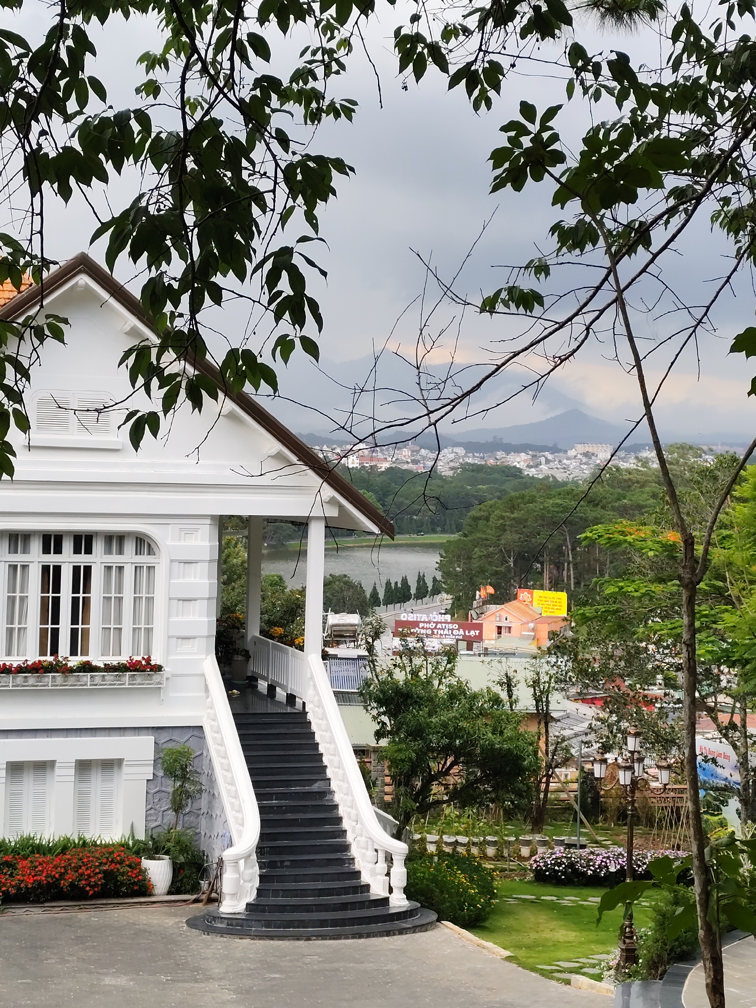 The image shows a white colonial-style house with a prominent dark staircase leading to a covered porch. The house has large windows and is surrounded by lush greenery and flowers. In the background, a lake is visible, with a city spread out on the hills beyond, and a hazy mountain range in the far distance. Several signs with Vietnamese text are visible in the mid-ground, indicating the location is likely a tourist area in Da Lat, Vietnam. The overall atmosphere is one of tranquility and natural beauty, characteristic of the city's landscape.