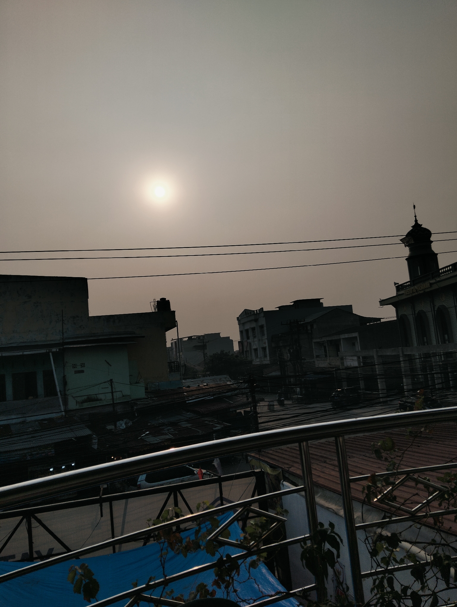 The image shows a view of the City of Medan, Indonesia, from a high vantage point, likely a balcony or rooftop. The sun is visible in the hazy sky, casting a diffused light over the scene. In the foreground, a metal railing with a pattern of interconnected geometric shapes and a blue tarp are visible, along with some leafy plants.
The middle ground features a collection of buildings, including what appears to be a mosque with a dome and minaret on the right side. Other structures include multi-story buildings with various architectural styles, some appearing residential and others commercial. Power lines crisscross the scene, indicative of an urban environment. The rooftops of some buildings are visible, some with corrugated metal and others with red tiles.
In the background, more urban structures fade into the hazy atmosphere. The overall impression is of a densely populated urban area in Medan, Indonesia, likely captured during daytime with low visibility due to haze or pollution. No people are clearly discernible in the image.