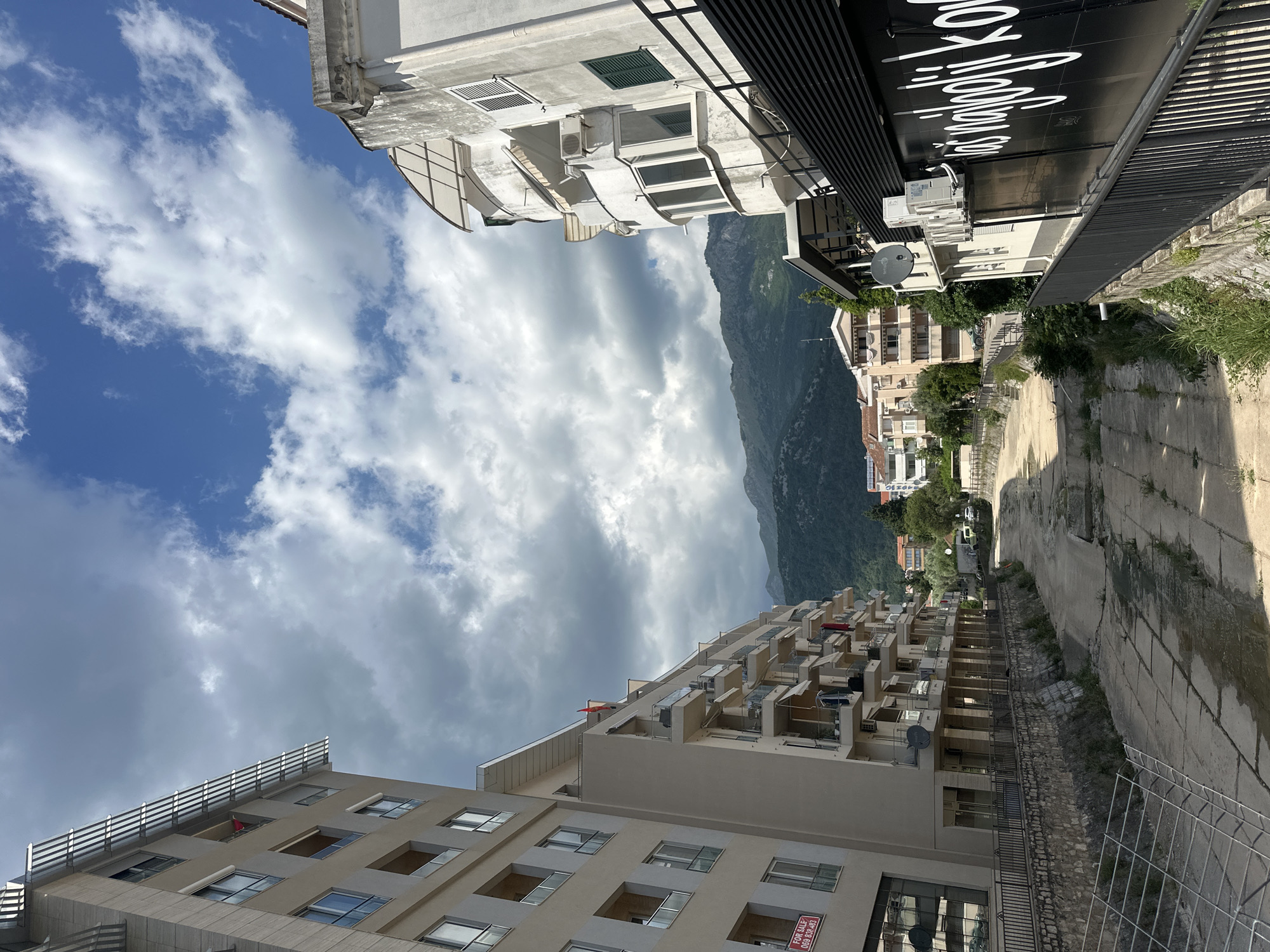 The image shows a street in Budva, Montenegro, framed by buildings on either side. A mountain range is visible in the background, partially obscured by clouds. The sky is a mix of blue and white clouds. The buildings are multi-story, with varying architectural styles and colors. A street runs between the buildings, constructed out of concrete. A metal wire fence is located along the right side of the street.