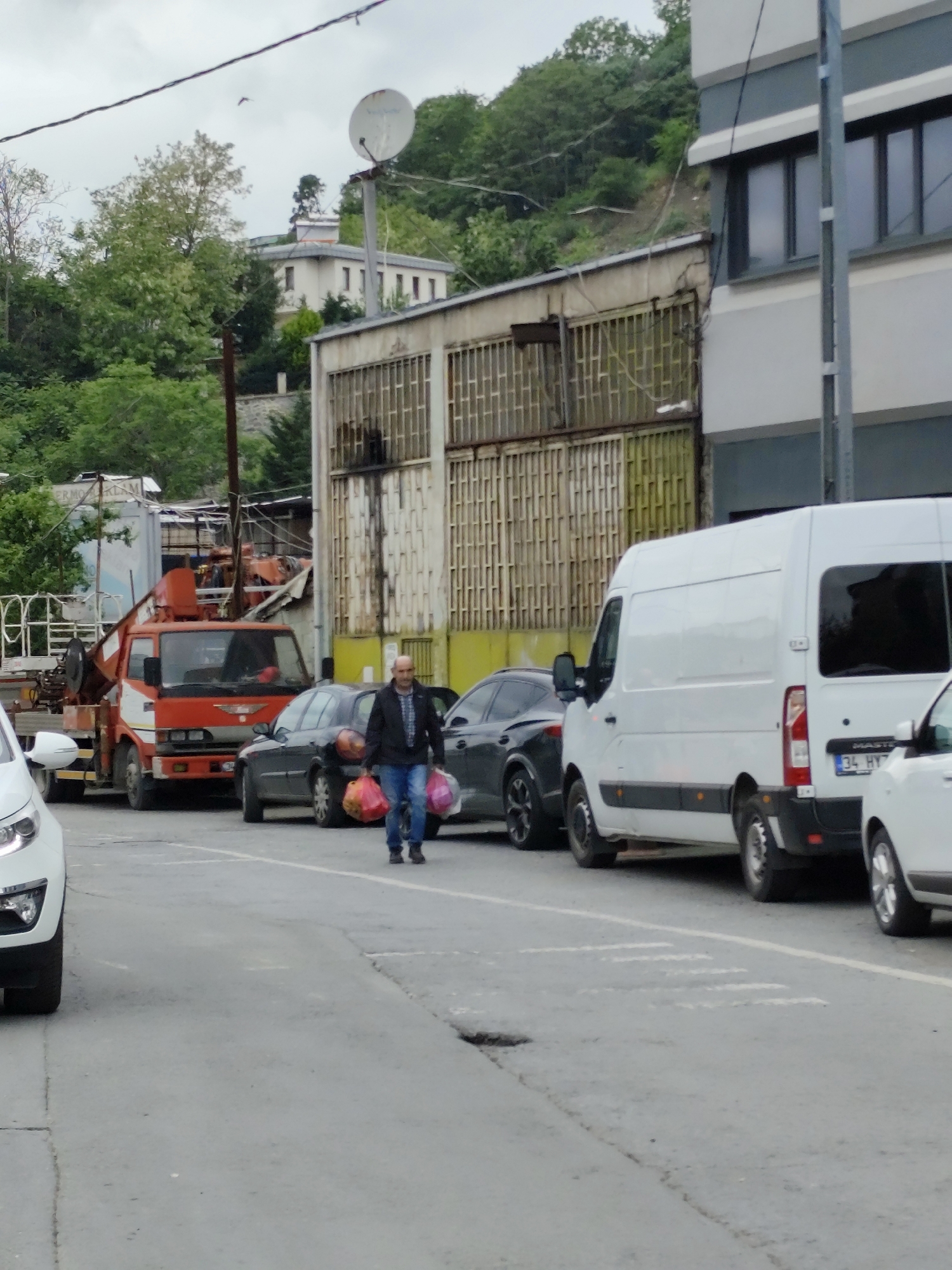 A street scene captures a middle-aged man carrying plastic bags, walking between parked vehicles in an urban environment. To the left, an orange boom truck with a white cab is parked, and a white van is positioned to the right. A building with a weathered facade stands in the background, adjacent to a more modern structure. Overhead, a satellite dish is mounted on a pole, with foliage-covered hills and another building visible in the distance. The street appears to be in Unknown, Unknown.