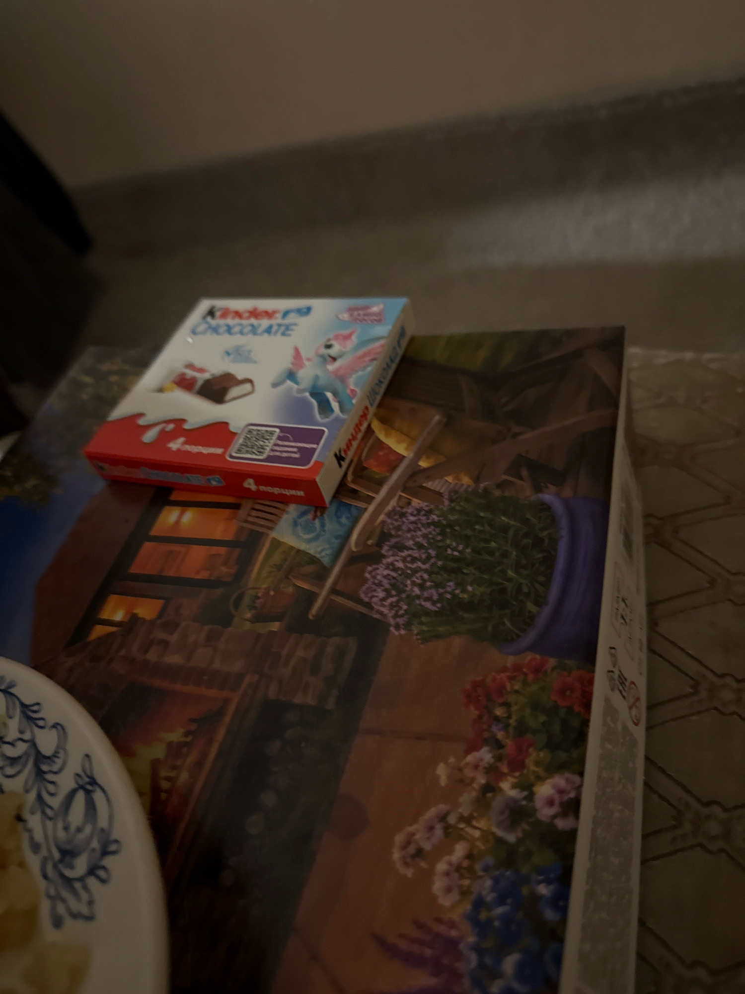 The image shows a close-up of a table surface. On the table, there is a box of Kinder Chocolate, which is light blue with red accents and features an image of a cartoon unicorn. The chocolate box is partially on top of a larger, flat object, possibly a puzzle box or book, with a printed image of a cozy, rustic interior scene featuring a fireplace and potted plants. In the foreground, a portion of a white plate with blue decorative patterns is visible, containing some yellow food items. The lighting is dim, suggesting an indoor setting, possibly in the city of Zavolzhye, Russia, based on the metadata.