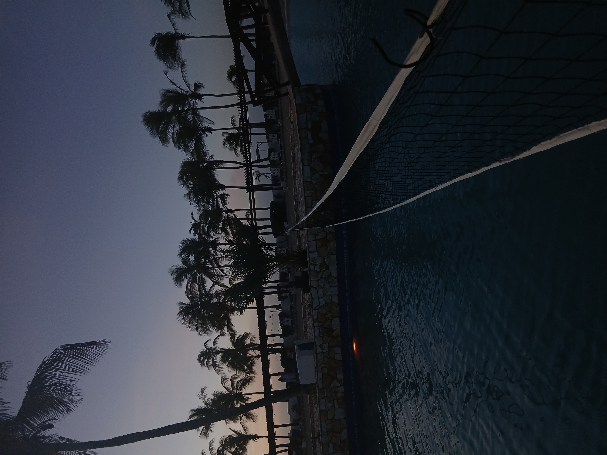 The image presents an outdoor recreational area, likely a resort or hotel, located within the city of Altagracia, Venezuela, captured during twilight. The foreground features a dark body of water, presumed to be a swimming pool, with a black volleyball net, outlined by white banding, stretched diagonally across its surface from the lower right. A single orange-yellow underwater light is visible within the pool, illuminating a small area near a multi-colored stone wall constructed from irregularly shaped blocks. Beyond this wall, a paved or sandy section contains several lounge chairs arranged beneath a wooden pergola-like structure. Numerous palm trees, uniformly silhouetted against the sky, are visible throughout the midground and background. One indistinct human figure, dressed in light-colored clothing, stands near the stone wall beneath the pergola, slightly left of the image's center. A dark wooden structure, possibly a bridge or walkway, extends over a portion of the water on the right side of the frame. The sky transitions from a lighter lavender-grey near the horizon to a darker indigo blue overhead.
