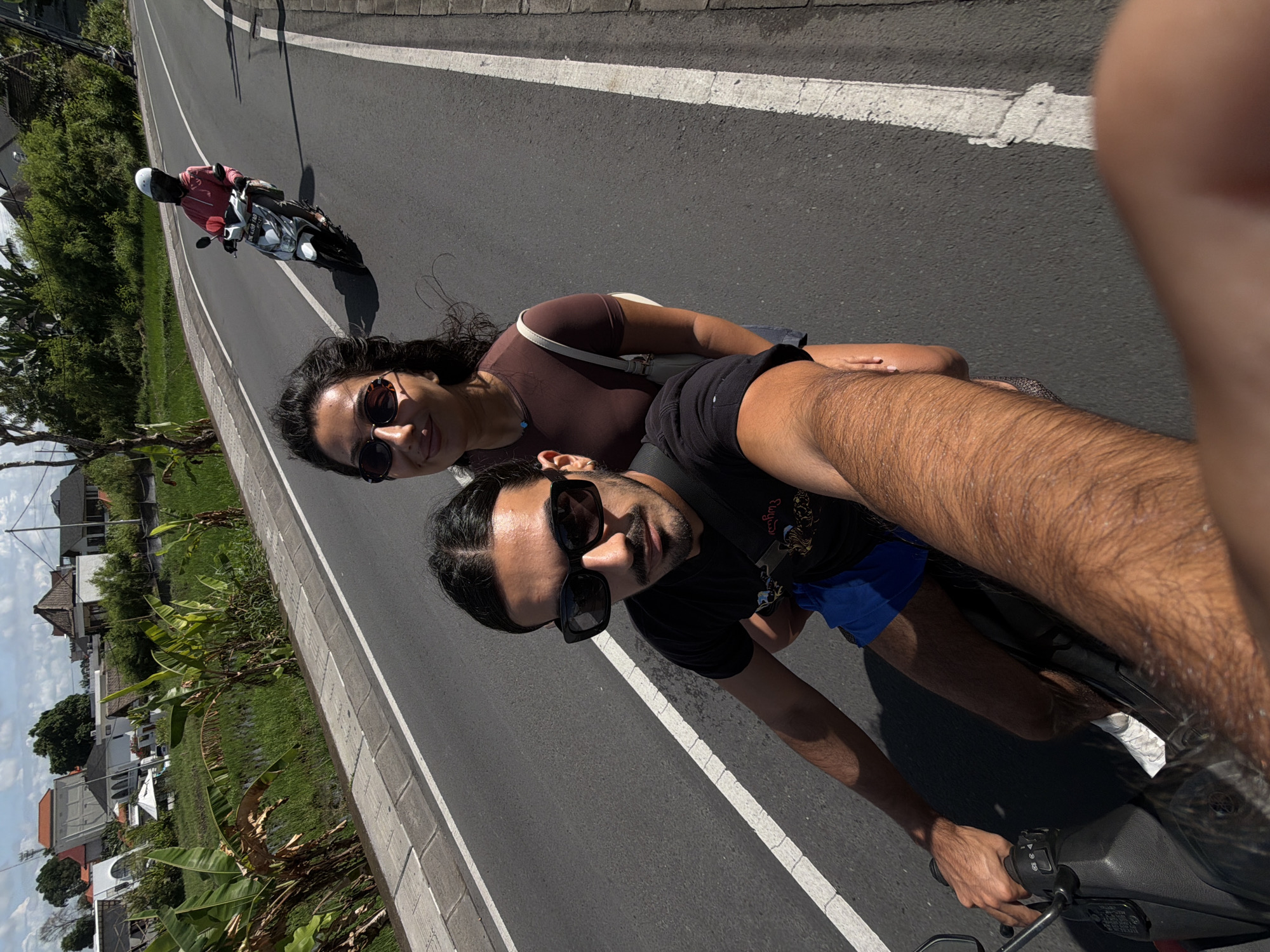 A tilted selfie-style image captures two people riding a scooter on a sunny day in North Kuta, Indonesia. The camera is held by the person driving, whose arm and torso are visible in the foreground. He has dark hair, a beard, and wears black sunglasses, a black t-shirt, and blue shorts. His left arm is extended to hold the camera, while his right hand grips the scooter's handlebar.
Behind the driver, a woman with dark, curly hair and sunglasses smiles towards the camera. She is wearing a brown top and has a white shoulder bag. Both individuals appear relaxed.
They are riding on a paved road with a solid white line marking the edge and a dashed white line separating lanes. In the background, another person on a white scooter is visible, wearing a white helmet and a red or pink shirt.
To the left of the road, there is a paved shoulder, followed by lush green vegetation, including banana plants, and what appears to be agricultural fields (possibly rice paddies). In the distance, several buildings with traditional Indonesian architectural elements are visible under a bright, partly cloudy sky. The strong sunlight casts distinct shadows, indicating midday. No visible text is discernible in the image.