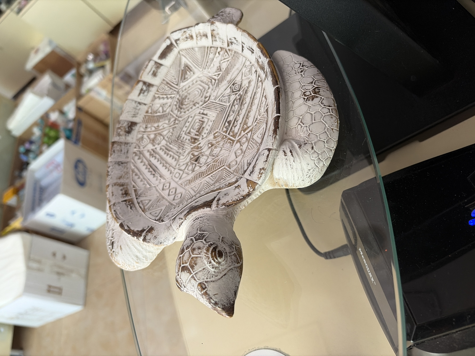 This image captures a decorative, white sea turtle statue with a distressed finish, resting on a clear glass surface. The statue is positioned such that its head faces the lower left, and its body extends towards the upper right, giving the impression the image is rotated 90 degrees counter-clockwise.
The turtle's shell is adorned with intricate, geometric, and tribal-like patterns carved into its surface. The white paint is chipped or worn in places, revealing a darker, brownish material beneath, which contributes to its aged aesthetic. The head and flippers of the turtle also exhibit a textured, scaly appearance with similar white and brown tones.
The clear glass surface reflects some elements of the room and shows minor imperfections like smudges or dust. Beneath the glass, on the right side of the image, a black electronic device is visible. The brand "MERCURY" is clearly inscribed on its side in white lettering, and a small blue light is also discernible on the device.
The background is out of focus but indicates an indoor setting. It contains several light-colored cardboard boxes and other miscellaneous items, suggesting a storage area or a somewhat cluttered room. The lighting is ambient, and no specific time of day can be determined. There are no people visible, and no activity or event is taking place, just a static display of the object. The location context provided is Sochi, Russia.