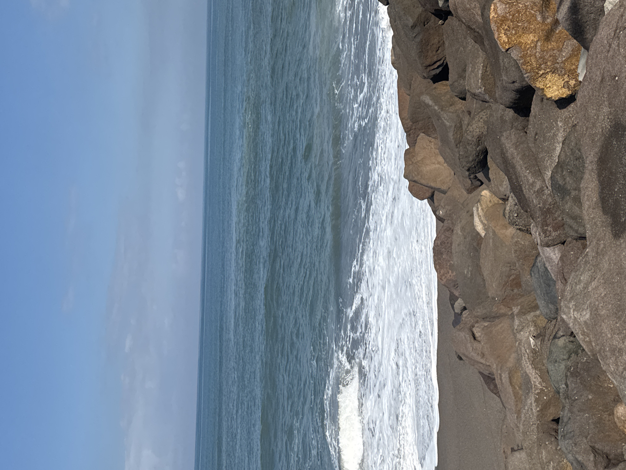 The image depicts a rocky beach with the ocean in the background. The foreground is dominated by a collection of large, irregularly shaped rocks of various brown and gray hues. The beach is composed of dark sand, and the ocean is a calm, blue expanse with small waves breaking near the shore, creating white foam. The sky above is a clear, pale blue with a few wispy white clouds. The horizon is visible where the sky meets the ocean. The scene is tranquil and natural.