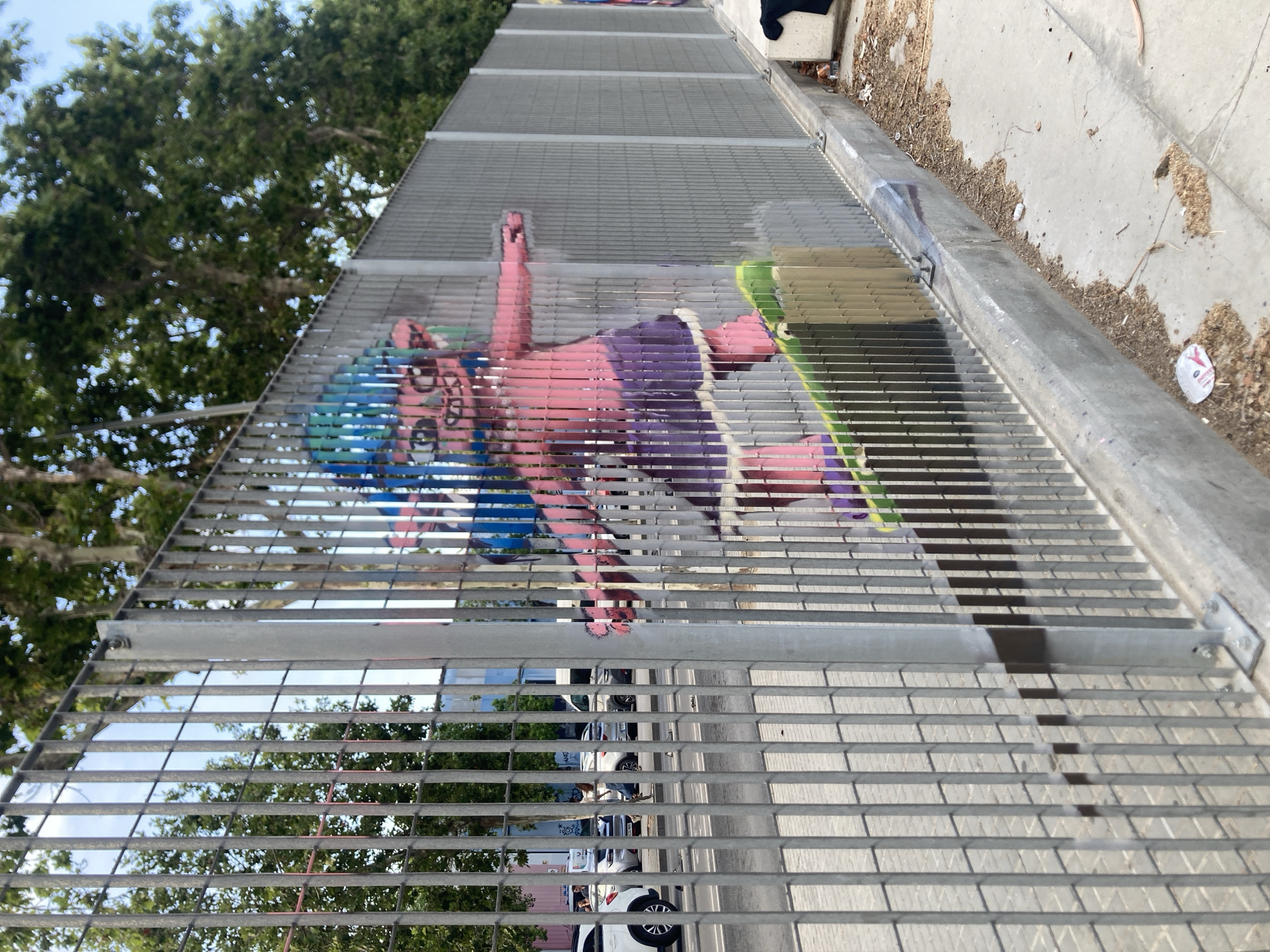 The image depicts a street scene in Badalona, Spain, likely in a commercial or industrial area, as suggested by the presence of parked cars and buildings in the background. A tall, grey metal fence with vertical slats runs across the foreground, obscuring a significant portion of the view. Through the fence, a vibrant, pink graffiti artwork of a cartoonish character with blue hair riding a green skateboard is visible. The character has a wide, grinning mouth and large eyes, and is depicted in a dynamic pose, as if performing a trick. The graffiti spans a considerable length of the fence. Beyond the fence, several vehicles are parked along a street, and lush green trees line the background, indicating an outdoor setting. The pavement in front of the fence is a mix of concrete and gravel, with some litter scattered about, including a white paper wrapper and wood shavings. The overall impression is one of urban street art contrasted with the everyday environment of a city.