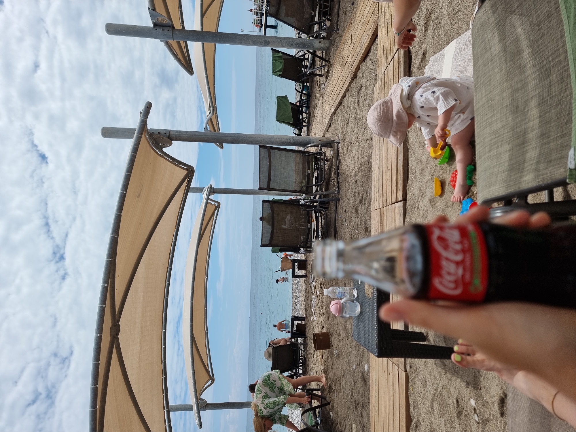 Here is a description of the image:
The image appears to be taken at a beach resort in Kemer, Turkey, judging by the beach setup (umbrellas, lounge chairs, wooden boardwalks) and the presence of Turkish lira water bottles. The sky is partly cloudy.
In the foreground, a person is holding a glass bottle of Coca-Cola. Their feet are visible, and they have lime green nail polish. A small black wicker table sits beside them.
To the right of the person is a child wearing a light-colored outfit and hat, playing with colorful plastic toys on the sand. An adult's arm reaches down to interact with the child.
The background shows the beach with lounge chairs, other people, and the sea. A pier-like structure is visible in the distance.