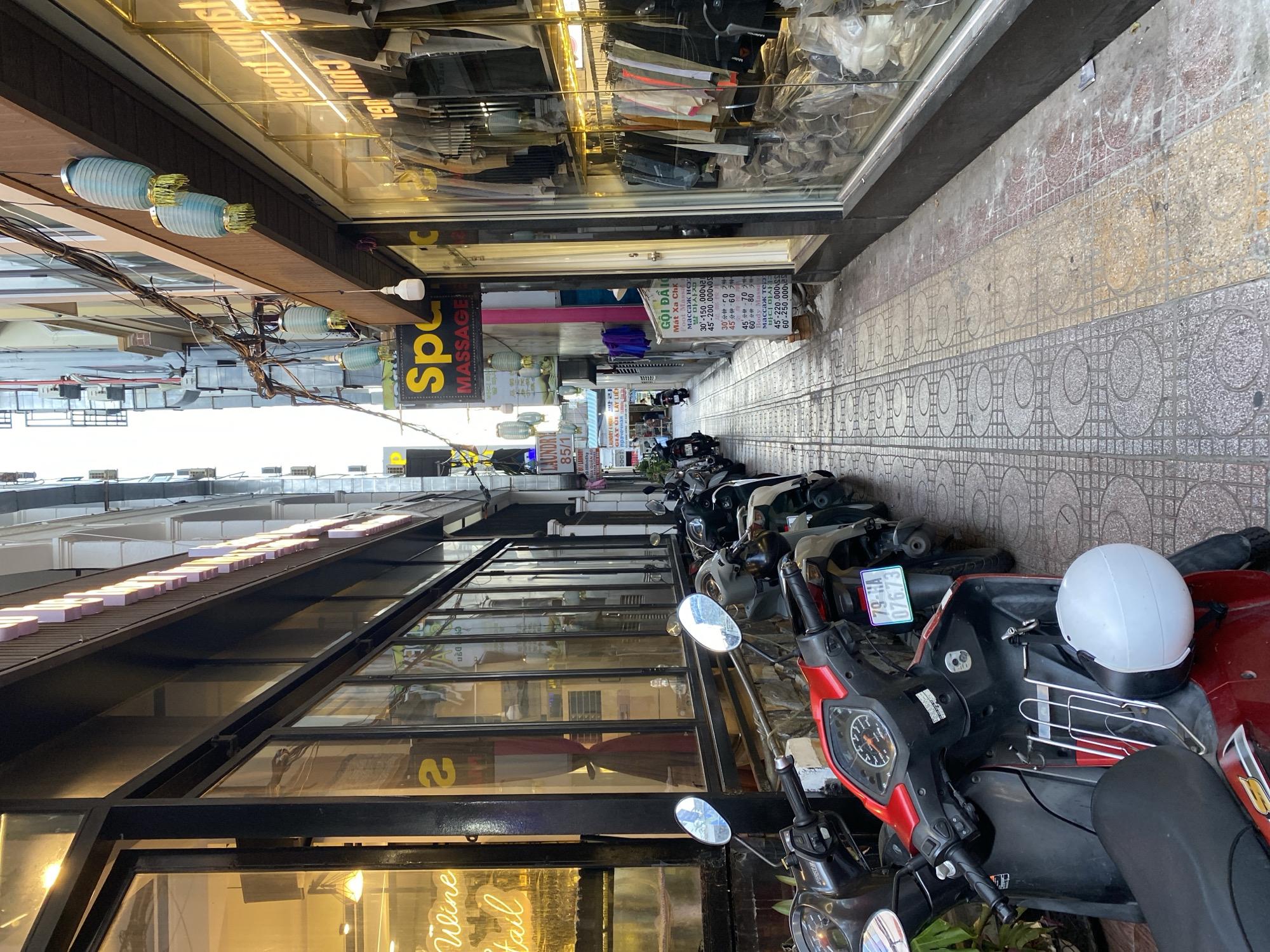 This is a street-level photograph taken in Nha Trang Ward, Vietnam, depicting a narrow alleyway lined with buildings on both sides. The alleyway appears to be a commercial area, with shops and businesses fronting the street. On the left, a shop with large glass windows displays merchandise and neon signage that reads "Wine Retail". Further down the alley, a laundry service with signage indicating "LAUNDRY 85/1" is visible.
On the right side of the alley, another shop with extensive glass windows showcases clothing racks filled with garments. A sign that reads "Spa Massage" is prominently displayed on a building in the center of the image. Several blue paper lanterns are hanging from wires strung across the alley.
The alleyway is paved with interlocking stone tiles, and a row of parked motorbikes lines the left side of the walkway, partially obstructing the view of the shops. A white helmet rests on the front of a red and black motorbike in the foreground. The overall impression is of a busy, urban environment with a mix of retail and service establishments. The lighting suggests it is daytime, with natural light illuminating the upper portions of the buildings and the alleyway itself, while shop interiors are artificially lit.