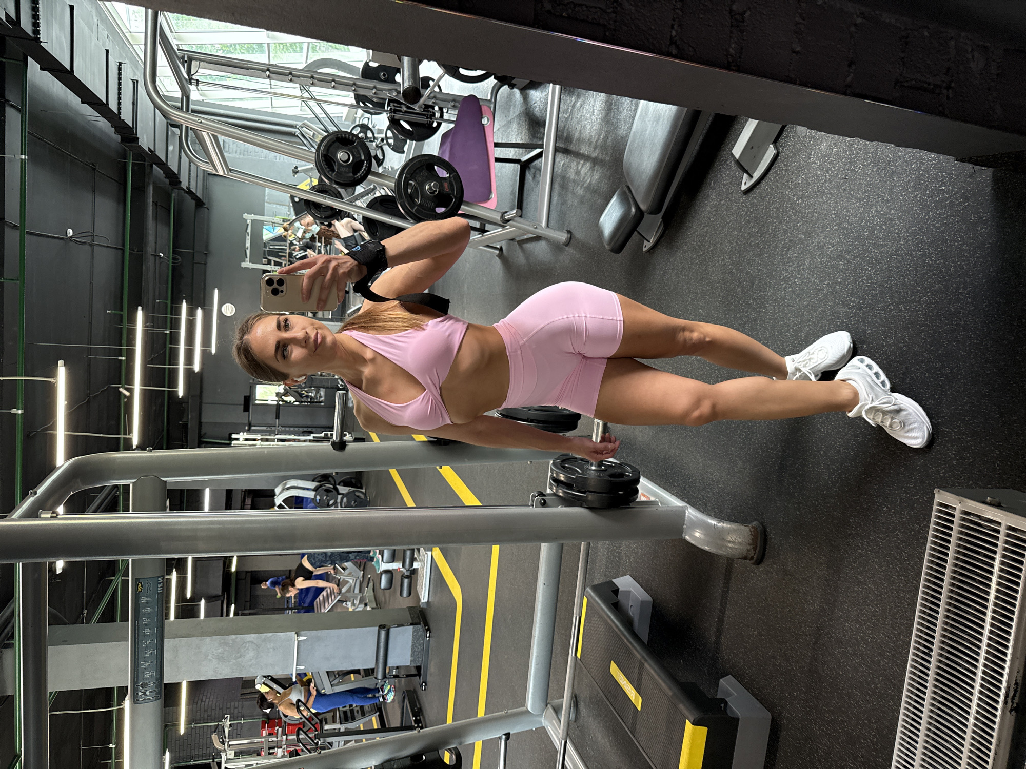 A full-body, selfie-style shot captures a woman in athletic wear posing in a gym. The woman, with long blonde hair, wears a light pink sports bra and matching shorts, paired with white sneakers. She holds a smartphone in her right hand, angled to take the photo, and has a black wristband on her left wrist. Her gaze is directed at the phone's screen.
The background reveals a well-equipped gym with various exercise machines, including a smith machine, weight racks with plates, a bench, and weight machines. Bright yellow lines mark pathways on the dark grey rubberized floor. In the mid-ground, other gym-goers are visible, some engaged in exercise. The environment suggests a modern fitness facility, likely in Saint Petersburg, Russia, given the provided context. The lighting is bright, originating from fluorescent tubes on the ceiling and natural light from windows visible in the distance. The overall impression is of a typical gym environment, with a focus on fitness and exercise.
