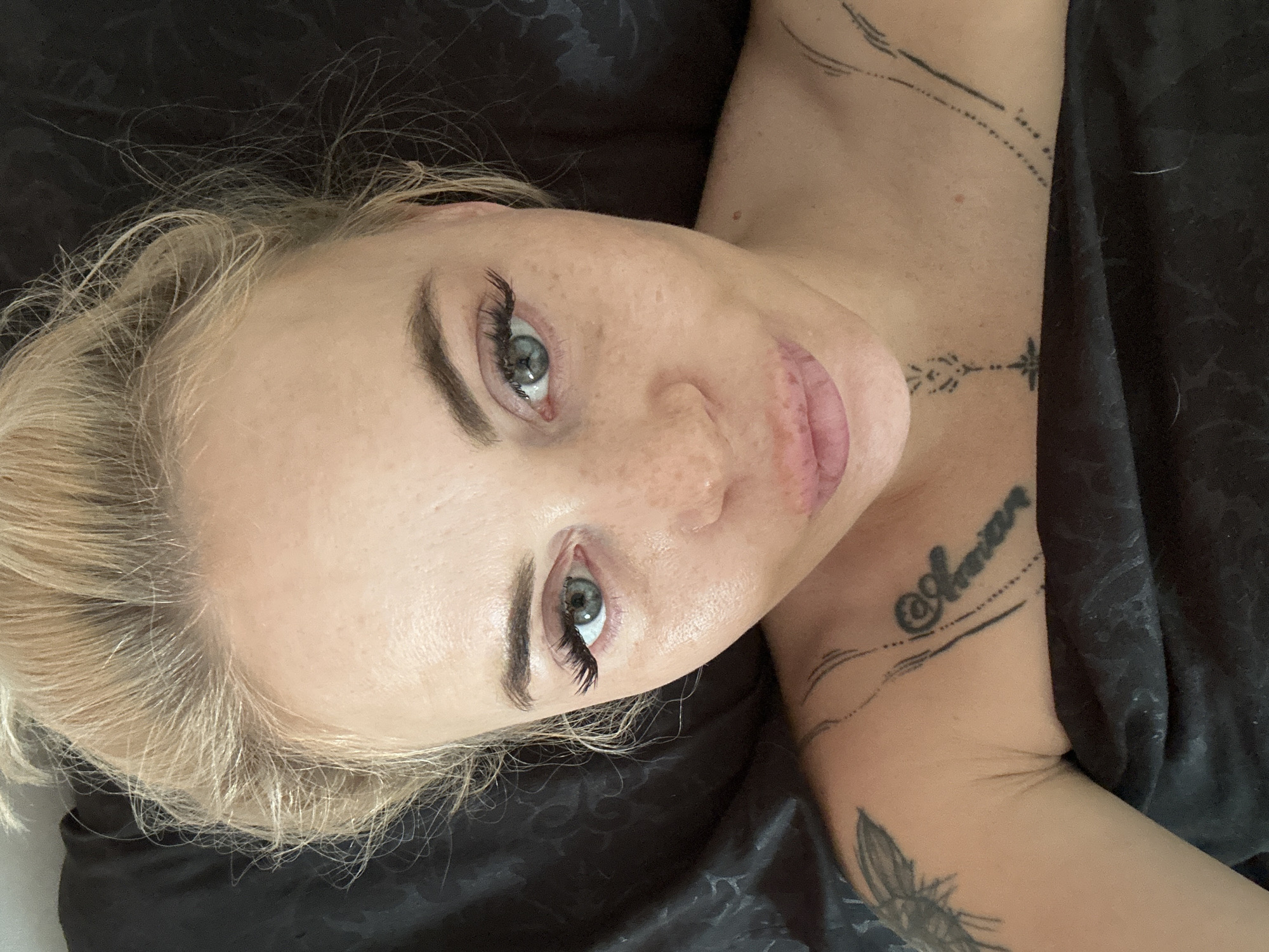 A close-up, overhead photograph depicts a fair-skinned adult female lying on dark bedding. Her blonde hair is pulled back loosely, with some strands framing her forehead. She has light-colored eyes, with voluminous false eyelashes; the upper lashes of her left eye (viewer's left) exhibit a subtle purple hue, while those on her right eye are black. Her eyebrows are dark and shaped. Freckles are visible across her nose and cheeks. Her lips are full and pinkish, formed into a slight pout. A small red blemish is present on her right cheekbone (viewer's right).
Multiple dark tattoos are visible on her upper body. On her left shoulder, a partial tattoo of an insect with wings is present. On her right chest/collarbone area, a script tattoo reads "@@Amaniar" (or similar), with a dotted line extending vertically below it, and abstract parallel lines above it. Another small geometric cross-like tattoo is located on her neck, below her chin.
The woman is covered by dark fabric, likely bedsheets or a blanket, which has a subtle, indistinct pattern. The background consists entirely of this dark fabric. The lighting is even and direct. The woman is looking directly at the camera. The location is unknown.