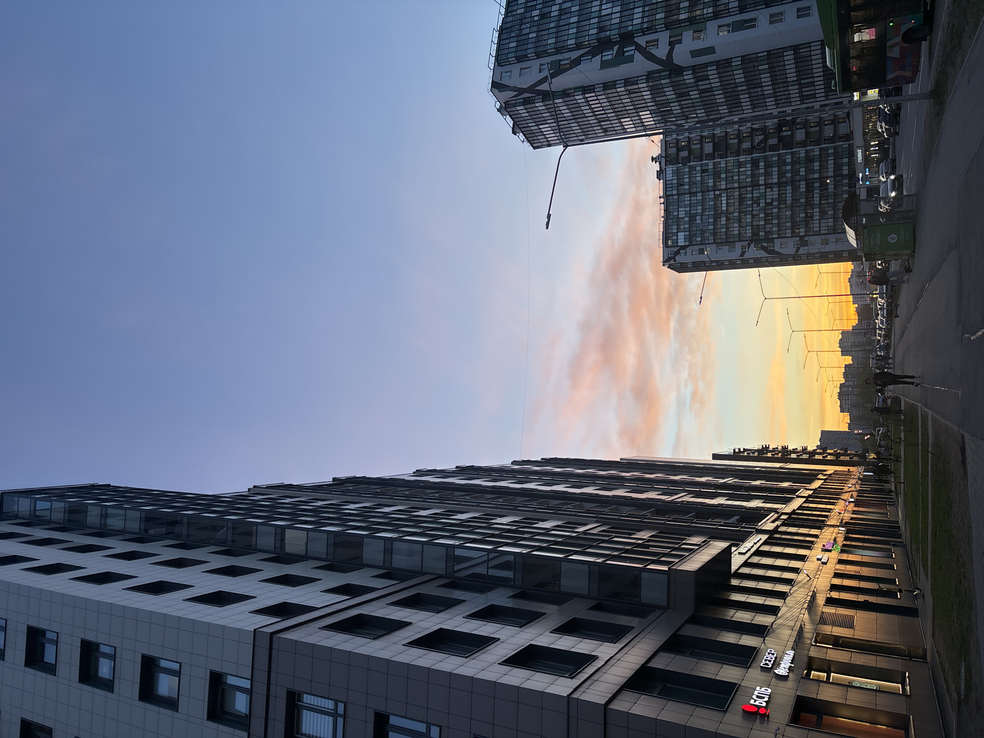 This image depicts an urban street scene in Kudrovo, Russia, at sunset. The sky transitions from a clear, soft blue on the left to warm, vibrant shades of orange, pink, and yellow on the right horizon, indicating the setting sun.
Two modern high-rise buildings dominate the scene. The building on the left features a dark and light panelled facade with numerous square and rectangular windows. The taller building on the right exhibits a more complex, patterned architectural style with extensive glazing.
A street runs along the right side of the image, populated by several vehicles, including parked cars and a green bus. A green dumpster is also visible near the road. A few pedestrians are seen walking on the sidewalk, their figures silhouetted against the bright sunset glow. A street lamp extends from the right-hand building over the road.
The golden light of the sunset bathes the right sides of the buildings and the distant city skyline, creating a warm and serene atmosphere. Visible text on the lower left building includes "БСПБ," "Север," and "Фармация," suggesting commercial establishments. The overall scene captures a tranquil moment in a contemporary urban environment at dusk.