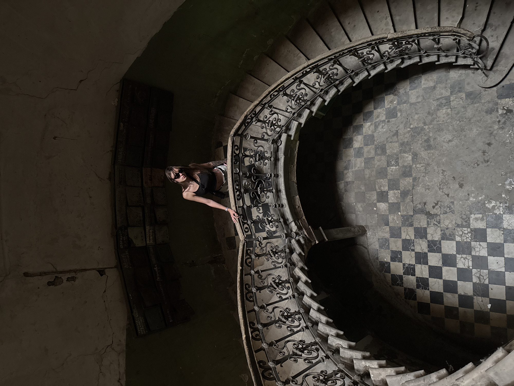 A young woman with sunglasses stands on a curved staircase in an old building. The staircase has a stone railing with ornate metalwork and stone steps. The floor is tiled in a black and white checkered pattern. Above the staircase, on the wall, are some inscriptions on what appear to be brickwork. The overall environment is dimly lit and shows signs of age and disrepair, with cracked plaster on the ceiling and walls. This scene is located in the abandoned Art Palace (State Museum of Georgian Folk Architecture and Applied Arts) in Tbilisi, Georgia, a location known for its historical and architectural significance.