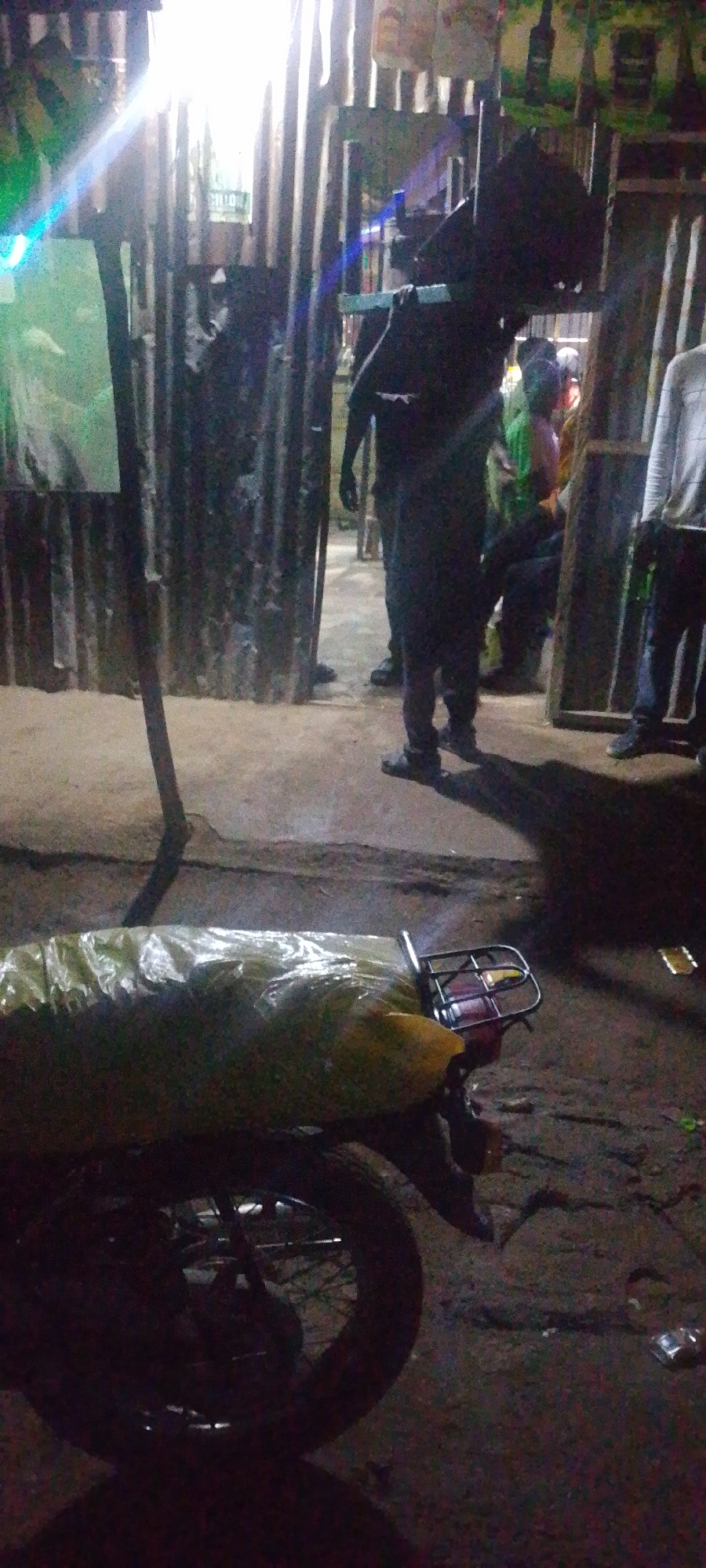The image depicts an outdoor scene at night, possibly in an urban setting in Unknown, Unknown. In the foreground, the rear of a motorcycle is visible, covered with a light-colored plastic sheet and resting on a rough ground surface. A black metal rack is attached to the back of the motorcycle. In the background, there is a corrugated metal structure serving as a wall or partition. Several individuals are present. One person, dressed in dark clothing, stands in the center, partially obscured by a black, amorphous object. Another person, wearing a light-colored shirt and dark pants, stands to the right. Other figures are visible in the background, further into the shadowed area. The scene is illuminated by artificial lighting, casting shadows on the ground. There are some posters or signs on the corrugated structure, and debris is scattered on the ground.