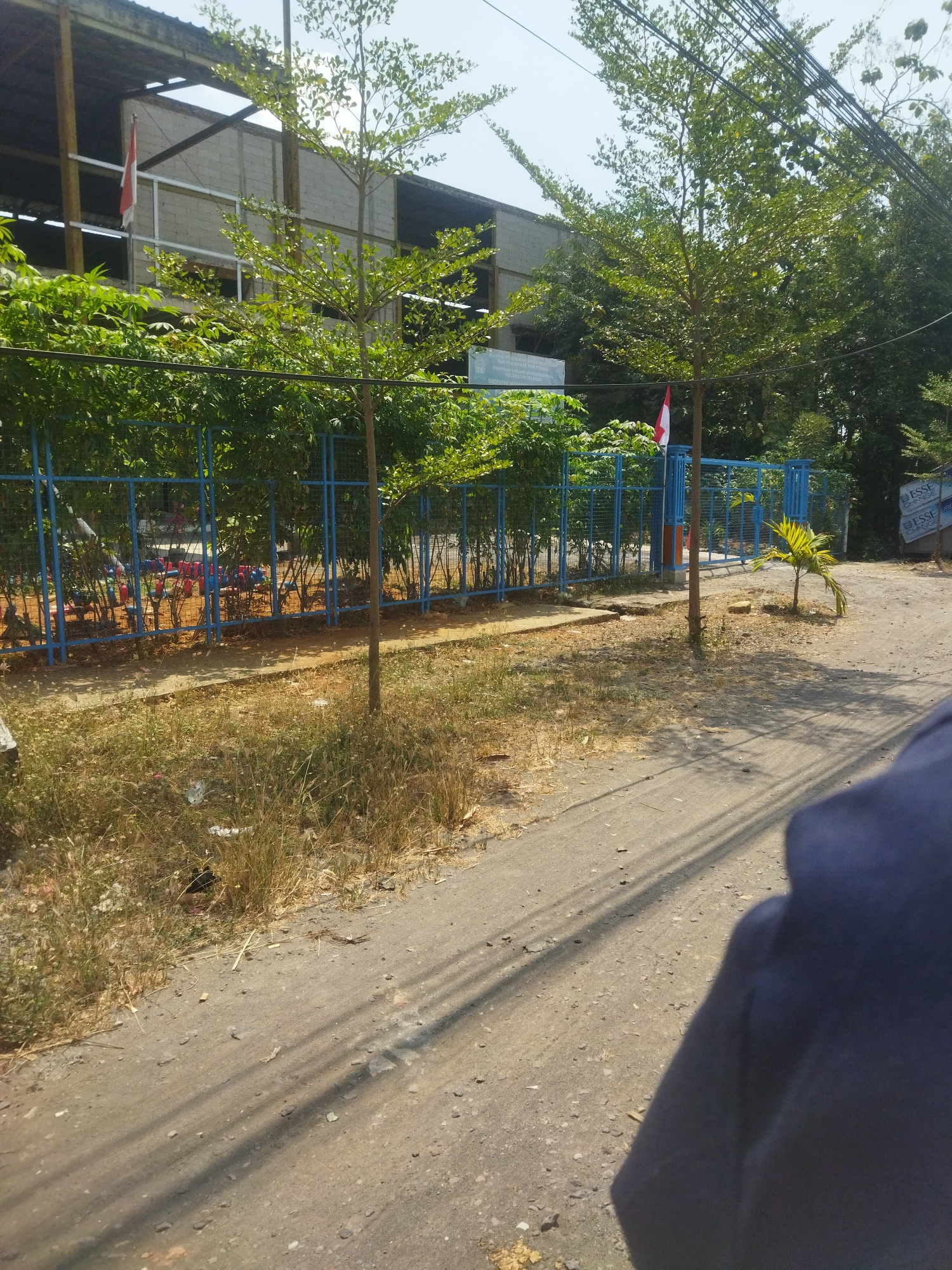 The image depicts a street scene in Nambangan, Indonesia, on a sunny day. In the foreground, a dirt road with sparse vegetation lines the side. A shadow of what appears to be a person wearing a dark blue garment is cast across the road from the right side of the frame. Further back, a blue metal fence encloses an area with various playground equipment, including red and blue structures, suggesting a children's play area. Behind the fence, lush green trees and bushes create a dense backdrop. To the left, a building with a grey concrete facade and a dark roof is visible, with an Indonesian flag displayed prominently. Two trees with sparse foliage stand in front of the fence. Further down the road, a small palm tree grows near the fence. In the far background, more trees and what appear to be posters with text are visible on a wall.