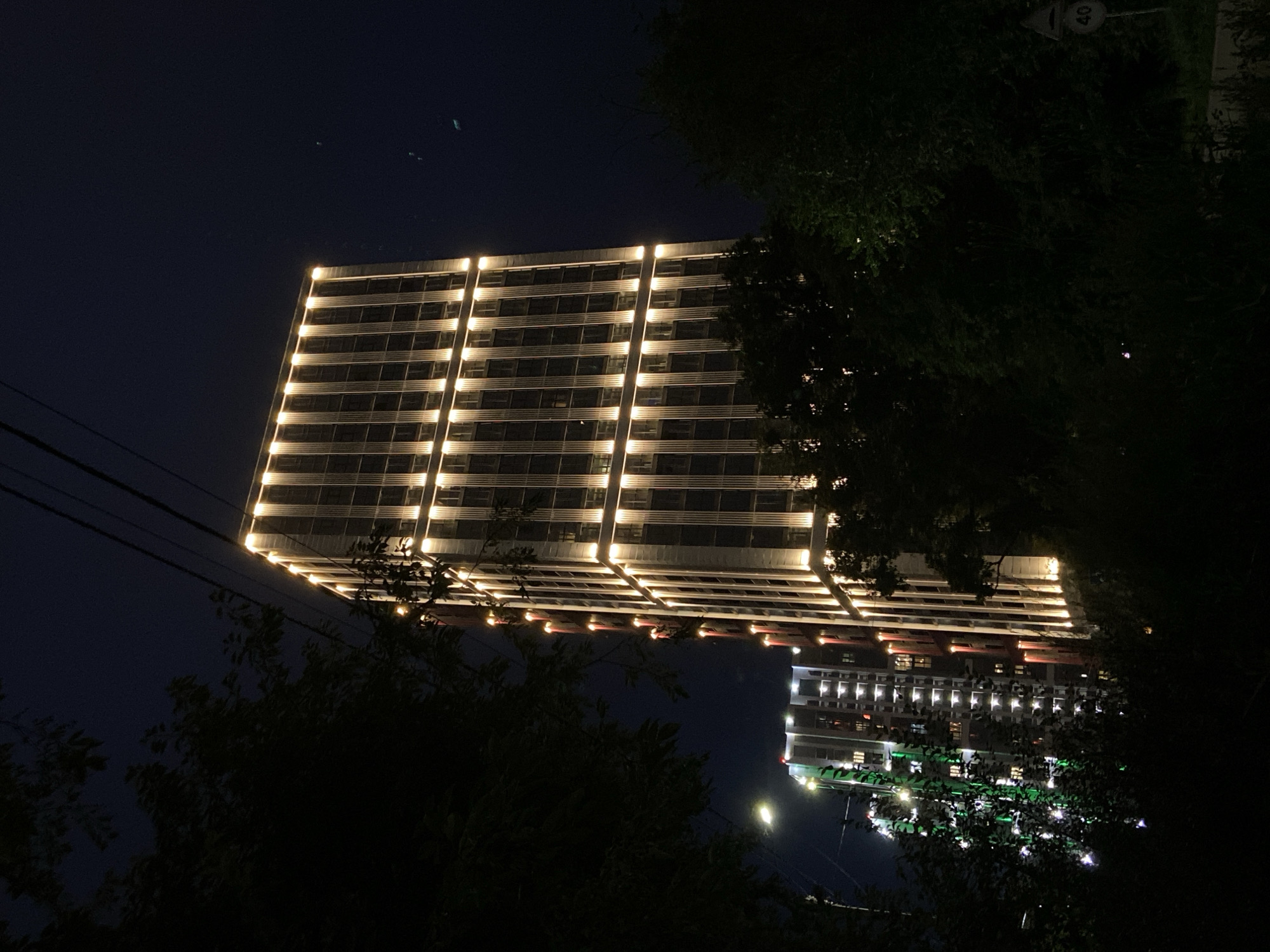 The image captures a nighttime urban scene in Blagoveshchensk, Russia, focusing on two illuminated high-rise buildings. The primary building on the right is a modern apartment complex with a facade adorned with vertical strips of warm white LED lighting, accentuating its architectural design. The building to the left, partially obscured by trees and greenery, is also a high-rise, lit with a combination of green and red lights, suggesting a different aesthetic or perhaps ongoing construction/illumination. The foreground is dominated by dark silhouettes of trees and foliage, framing the illuminated structures against the deep blue of the night sky. Overhead, electrical wires are visible, and a traffic sign indicating a speed limit of 40 is present on the right side of the image. The scene conveys a sense of urban development and contemporary nighttime lighting. There are no people or animals visible in the image.