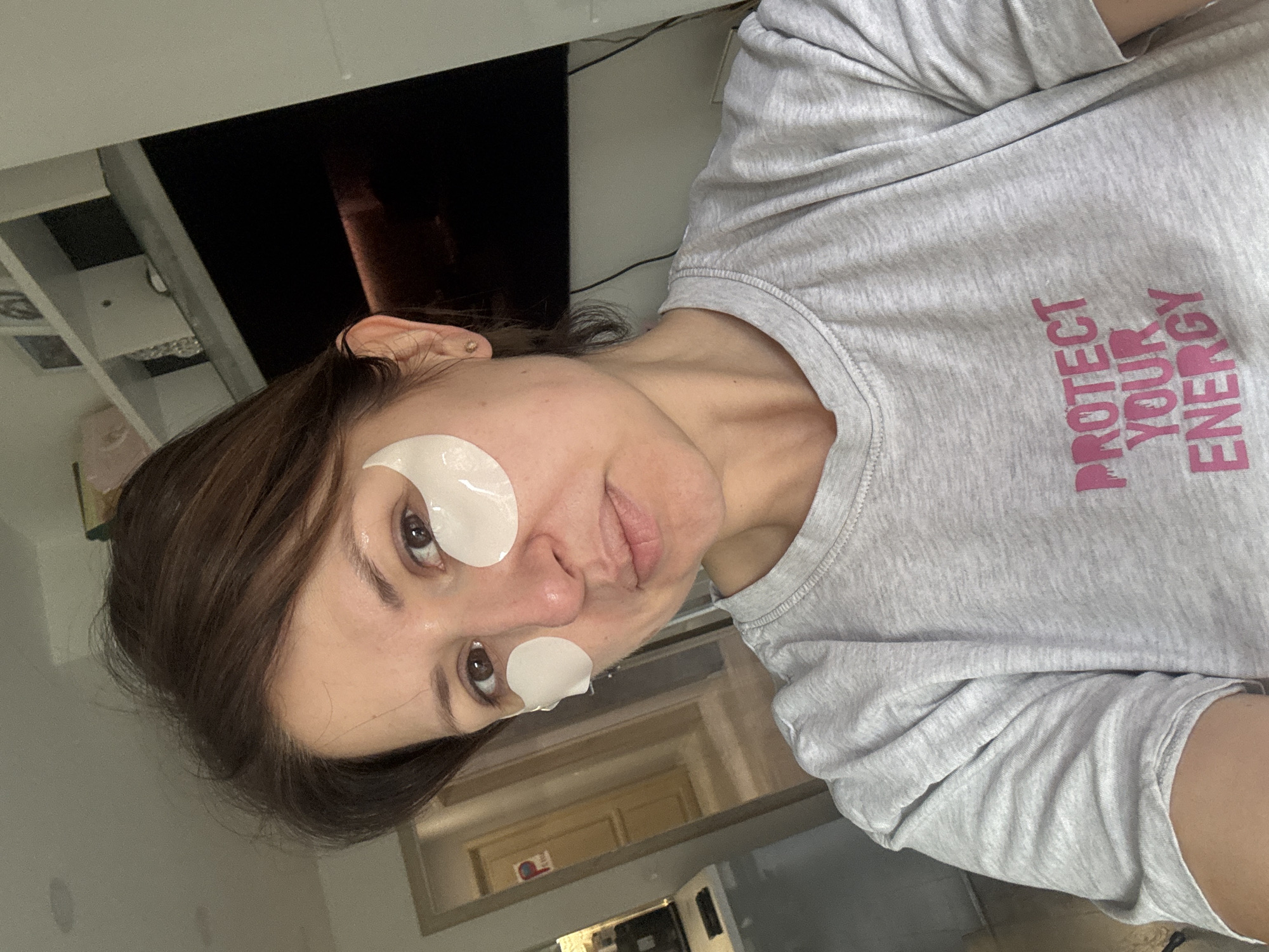 A woman is seen indoors, possibly in an apartment or house. She is wearing a light gray t-shirt with the phrase "PROTECT YOUR ENERGY" printed on it in a light pink color. The woman has two white circular facial patches applied to her face, one under each eye.
The background shows a room with a doorway and what appears to be a television or dark screen mounted on the wall. The setting does not immediately suggest any specific location in Sofia, Bulgaria. The scene depicts a casual domestic setting.