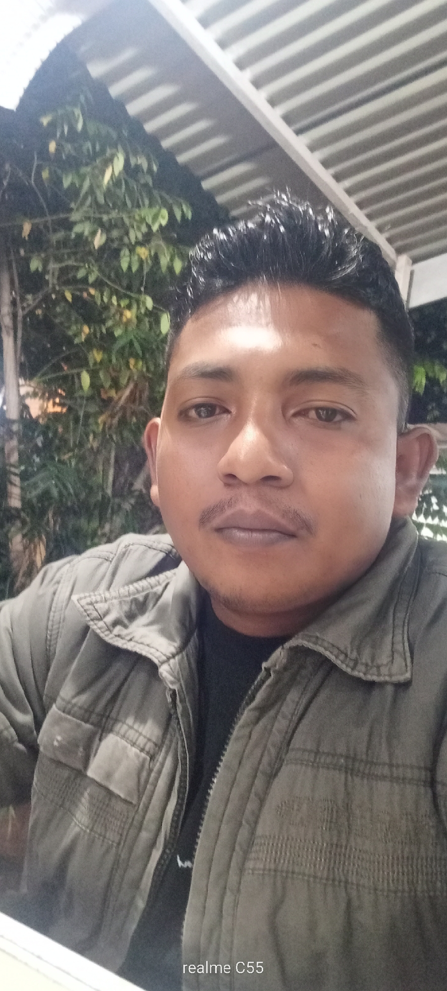 This close-up portrait, likely a selfie, features a man of Southeast Asian descent looking directly at the camera with a neutral expression. He has short, dark, textured hair and a thin mustache, with faint stubble visible on his chin. He is wearing a dark, possibly olive green or grey, zip-up jacket over a dark t-shirt.
The setting appears to be an outdoor or semi-outdoor area, likely at night, in Lhokseumawe, Indonesia. Artificial lighting illuminates the scene, casting highlights on the man's hair and jacket. Behind him, dimly lit green foliage and tree branches suggest a natural backdrop. Above, a prominent light-colored, slatted or corrugated roof structure indicates a covered space, possibly a patio, cafe, or restaurant.
No specific activity is depicted other than the man posing for the photograph. The overall mood is casual and direct. The text "realme C55" is visible as a watermark in the bottom right corner of the image.