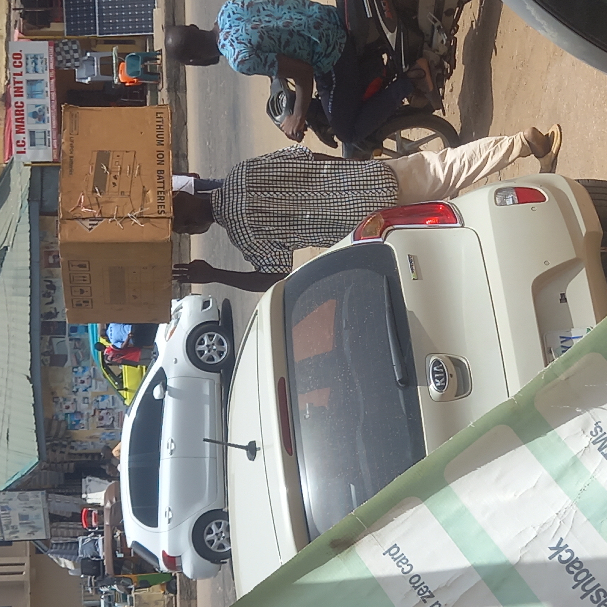 The image captures a scene in Dadi Riba, Nigeria, featuring a man in a checkered shirt and light-colored pants carrying a box labeled "LITHIUM ION BATTERIES" near a white Kia car. Another individual is seated on a motorcycle nearby, wearing a blue patterned shirt. The setting appears to be an urban street, indicated by the presence of parked cars, a motorcycle, and storefronts with signs, including one that reads "I.C. MARC INT'L CO." Solar panels are visible on top of a building. The scene suggests a transaction or delivery of goods taking place in a busy commercial area.