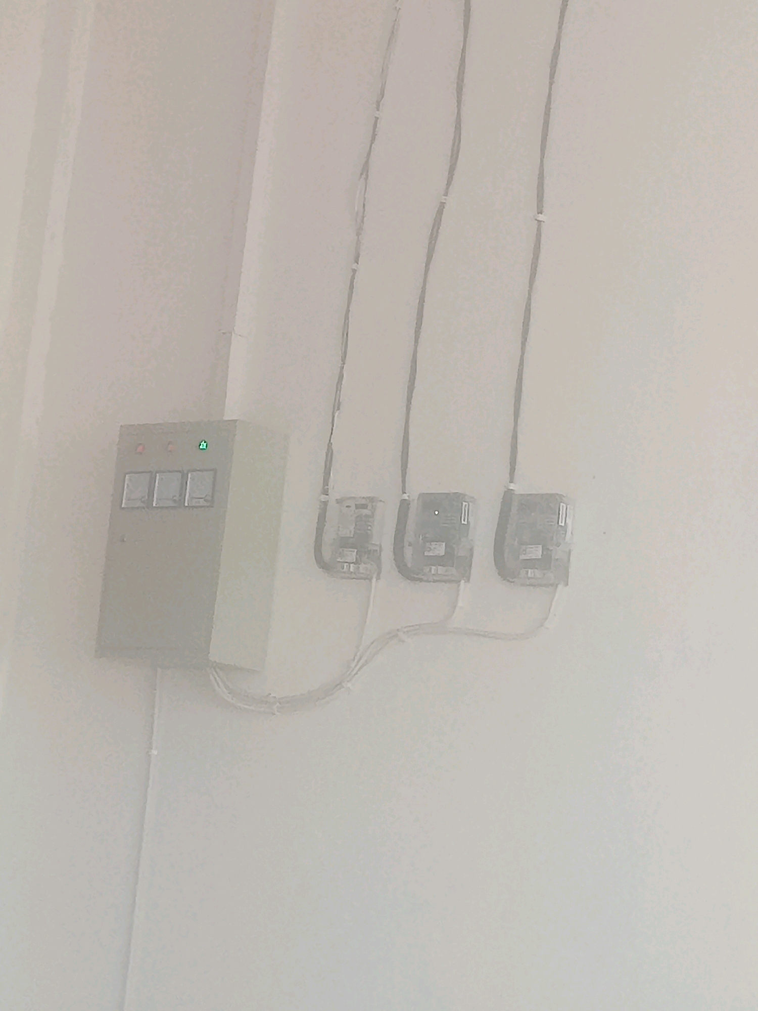 The image depicts an electrical panel and three meters mounted on a white wall. The electrical panel is a light-colored box with three analog meters on its front, above which are three indicator lights, two red and one green. To the right of the panel, three smaller, dark-colored electrical meters are mounted on the wall, each connected by cables. The meters are arranged in a row, and the cables are secured to the wall with white clips. The overall environment appears to be indoors, likely in a utility area or hallway, with a light-colored floor visible at the bottom of the frame. The scene does not contain any people or evidence of specific activities.
