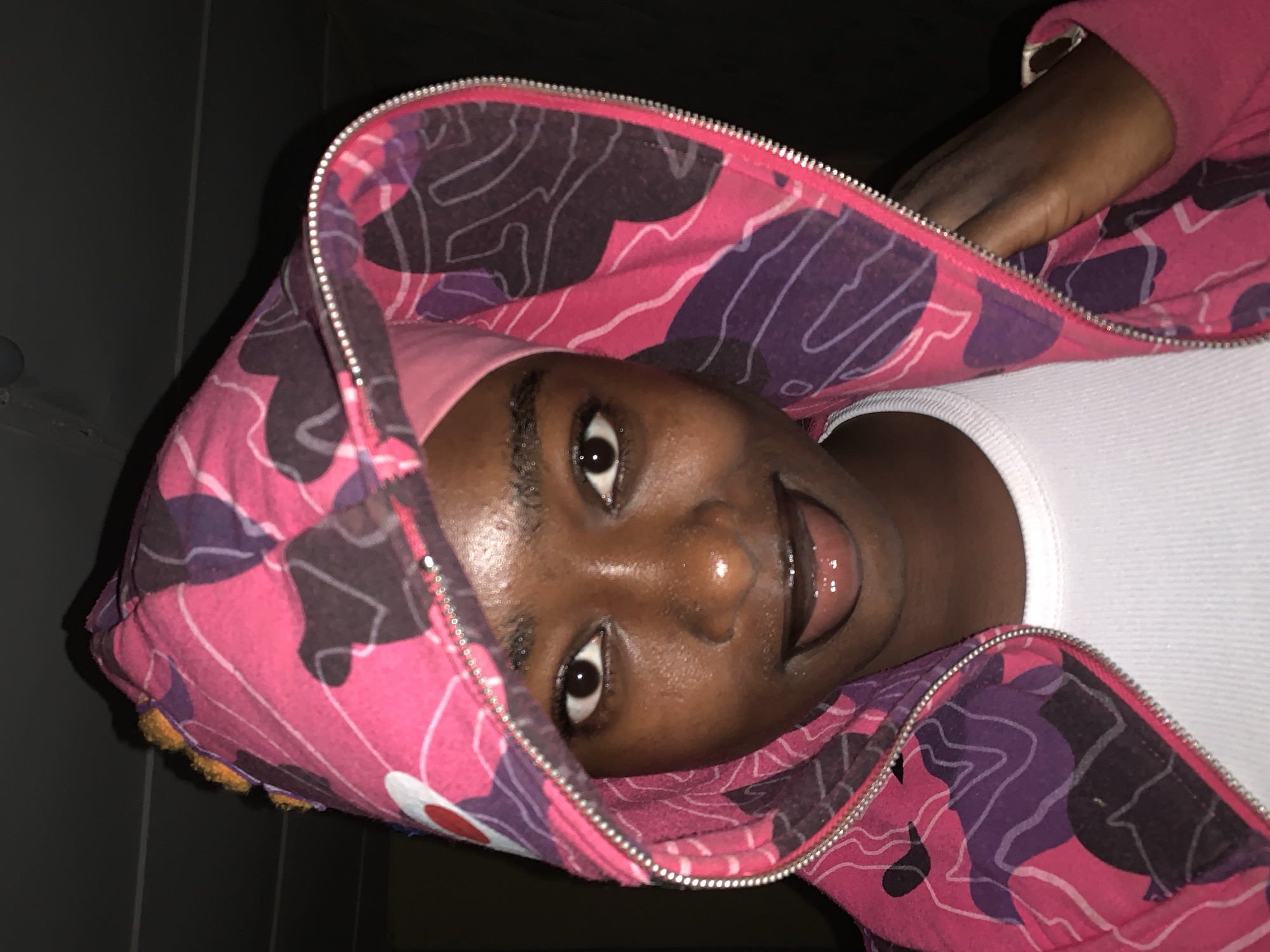 A medium close-up shot depicts a Black person wearing a pink and purple camouflage hoodie and a white undershirt. The person's face is centered and angled towards the viewer, with eyes open, lips slightly parted, and visible teeth. The hoodie's zipper is partially open. A hand is visible partially within the hood. The background appears to be a dark wall. The image doesn't provide information related to Rafin Par, Nigeria, nor does it provide information enabling the scene to be located.