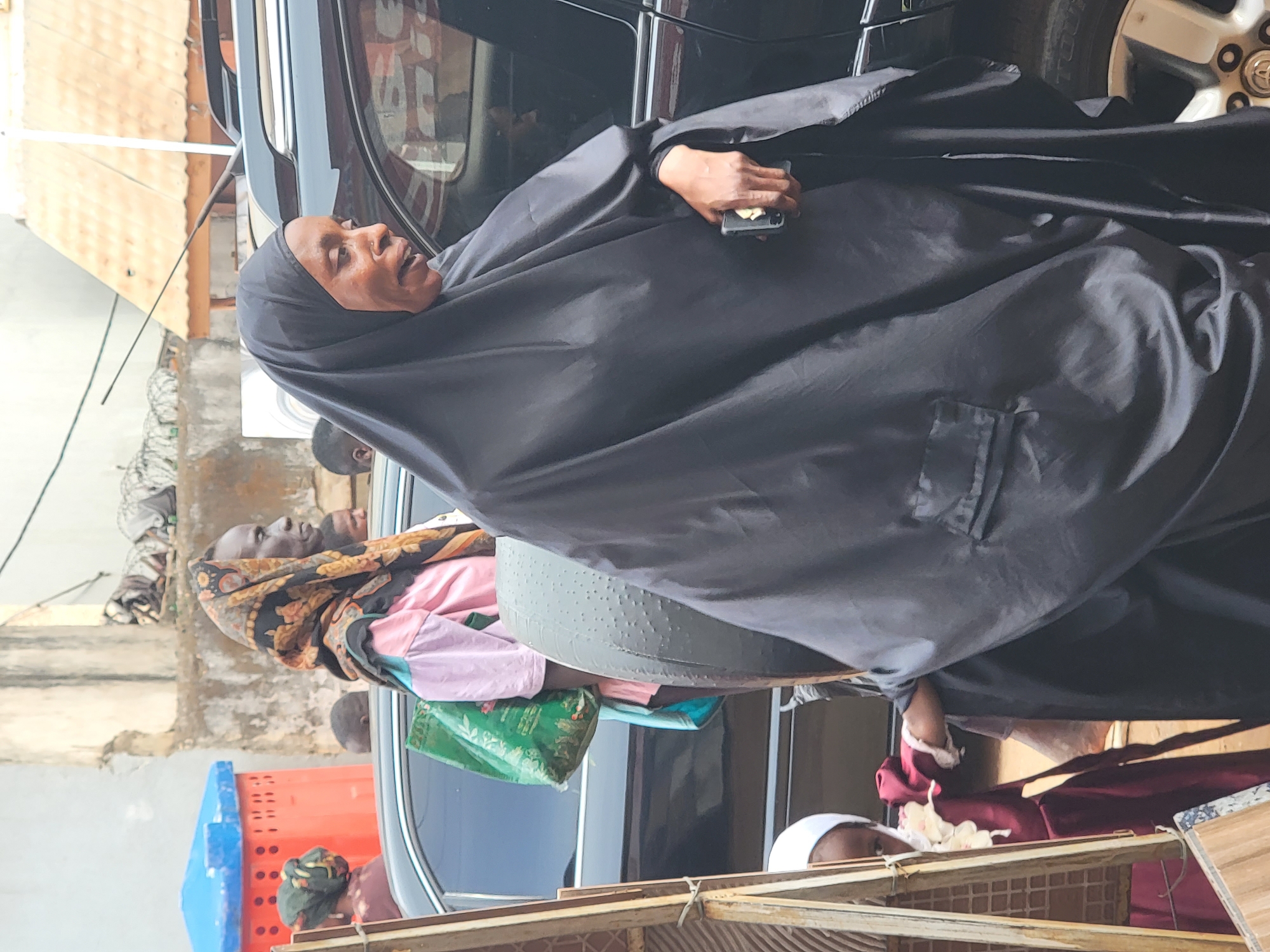A medium shot depicts an adult female in Maiburiji, Nigeria, wearing a black hijab while holding a smartphone. She stands beside a black vehicle. Another woman with a patterned headscarf and pink clothing stands near her, holding a collection of bags. An assortment of items including a blue crate and a green bag is visible. The setting appears to be outdoors near a building and a partial wooden structure.