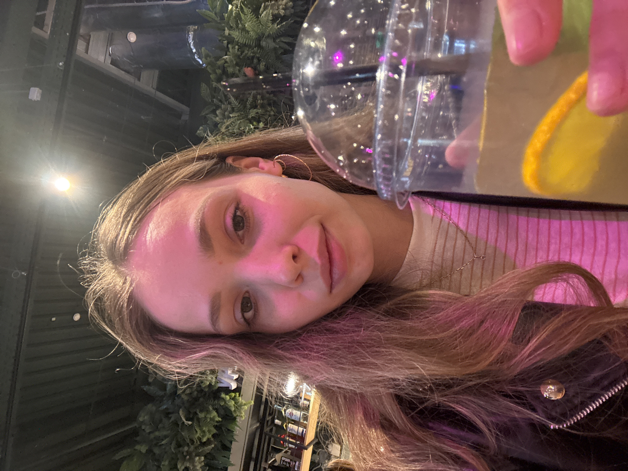 A young woman with long, light brown hair is holding a plastic cup containing a beverage. The beverage appears to contain a slice of citrus fruit. The woman is wearing a light-colored striped shirt and a black jacket with a zipper. In the background, there are plants and shelves with bottles on them. The lighting is pink.