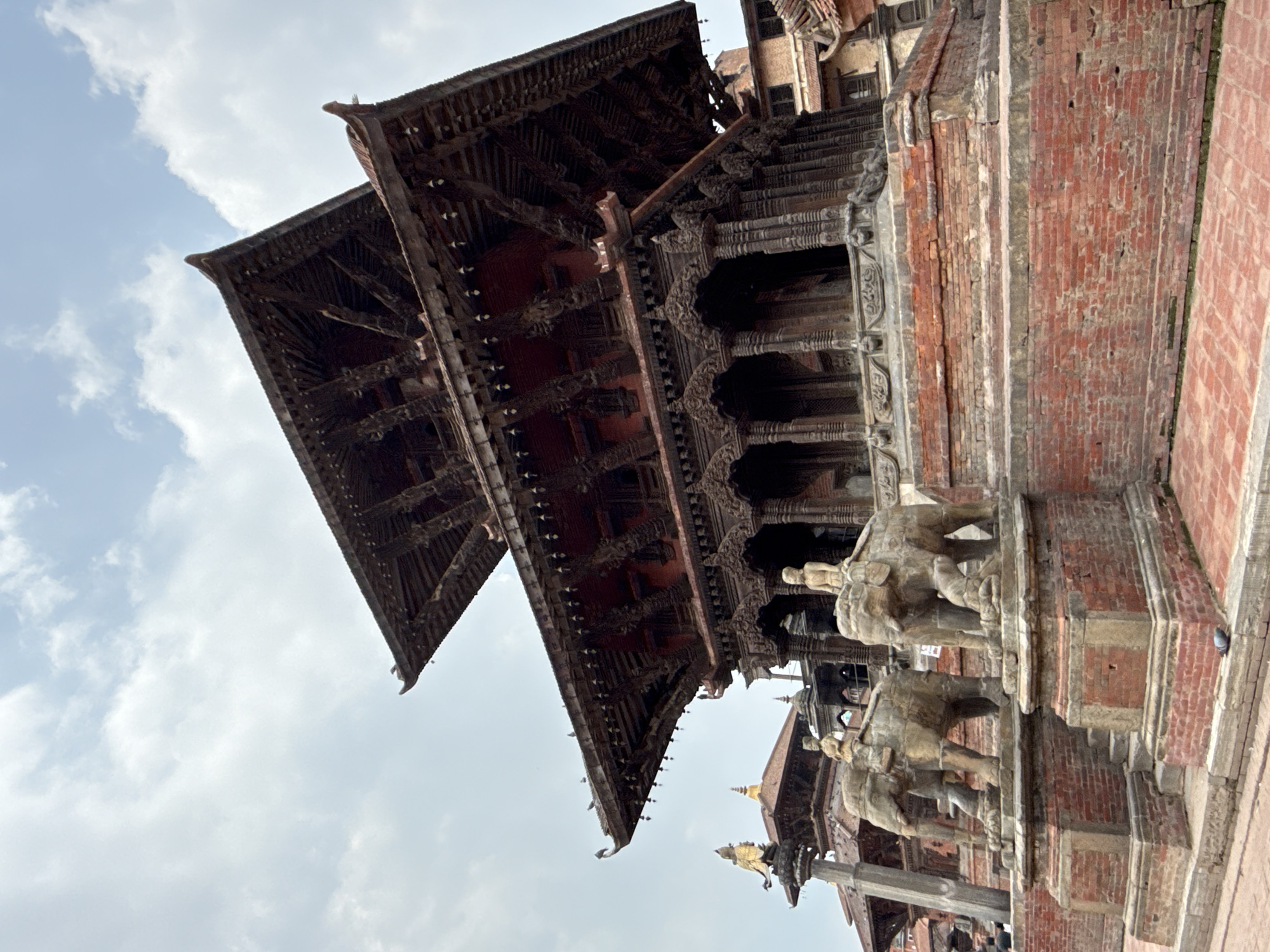 The image shows a temple structure in Patan Durbar Square in Lalitpur, Nepal. The temple has a multi-tiered wooden roof intricately carved. Below the roofline, there are rows of columns and other architectural details. To the right, there are brick structures with brickwork patterns. In the foreground, there are stone or concrete elephant statues. The sky behind the temple is pale with cloudy patches. The photo appears to have been taken from a low angle, looking upward at the building.