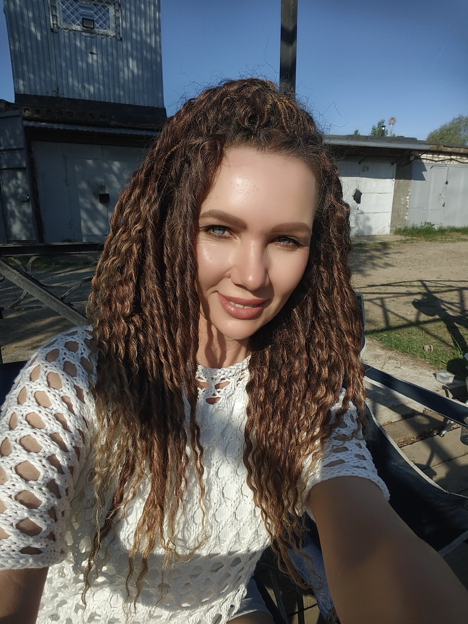 A medium shot depicts a Caucasian woman with long, brown curly hair, partially styled up, and blue eyes, wearing a white crocheted top. She is smiling slightly and appears to be taking a selfie.
The background includes a metal garage structure to the left and a building similar to garages in the back right, suggesting an urban industrial area or possibly the outskirts of a city. The area seems to be a sunny outdoor location with some foliage visible. The city location is Unknown, Unknown.