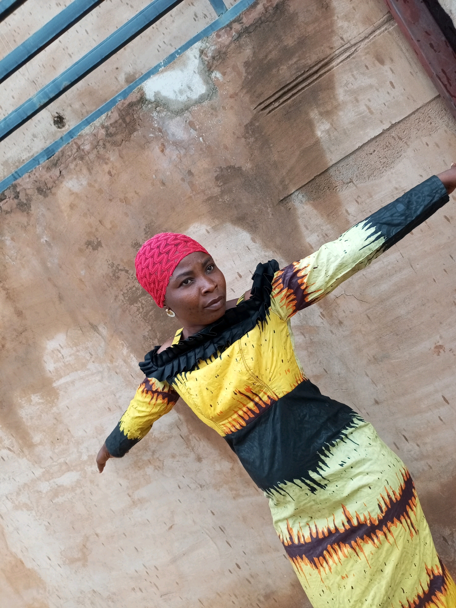 An angled shot shows a woman wearing a vibrant yellow and black patterned dress and a red headscarf. She stands outdoors against a textured, tan-colored wall, with blue metal bars visible at the top left of the frame. Her left arm is extended towards the right, and her right arm is bent and slightly raised. The wall appears weathered, with patches of lighter color and some rough textures. The lighting suggests it is daytime.