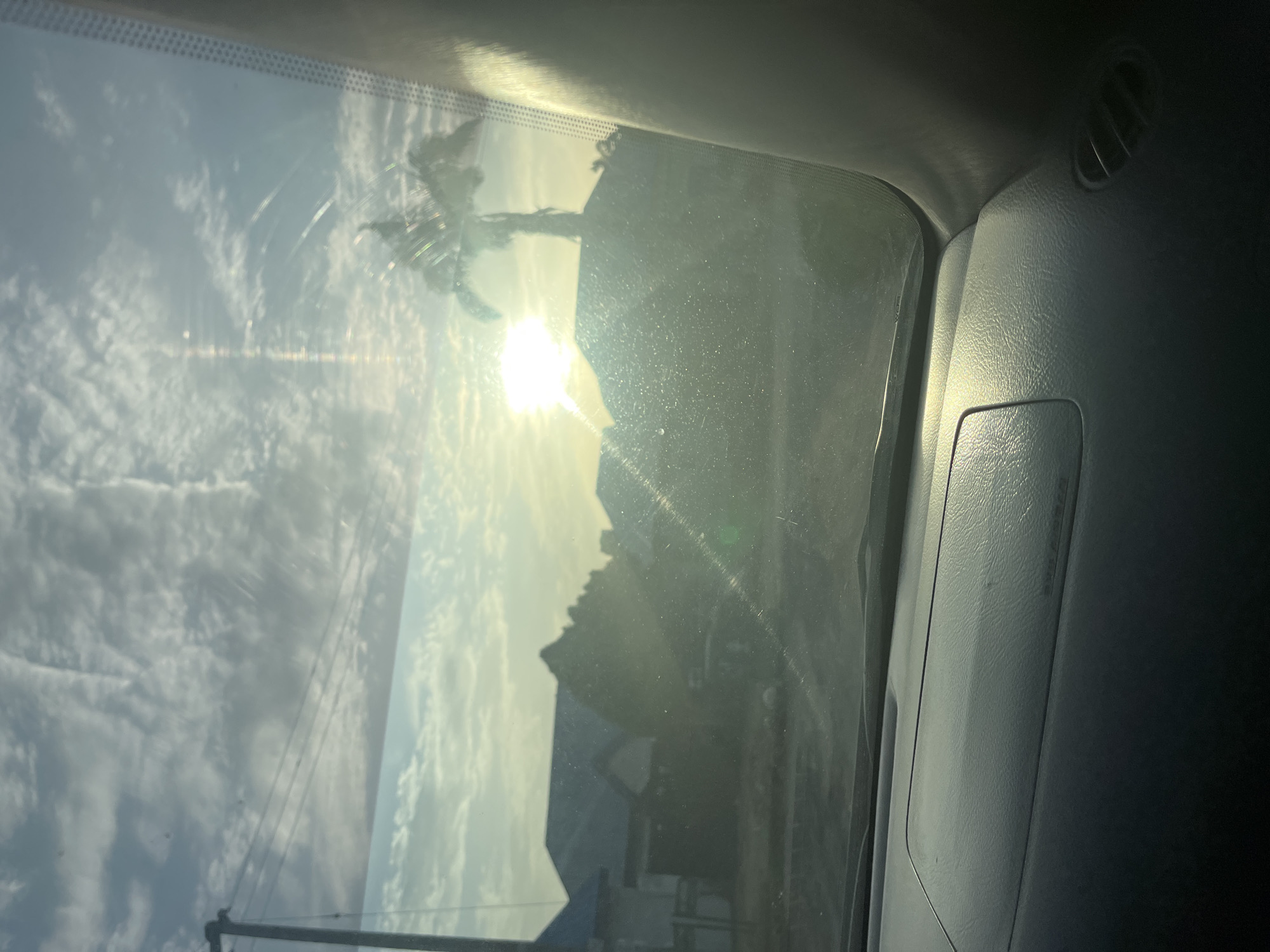 The image, taken from inside a vehicle, shows an obstructed view of a street scene in what may be Abuja, Nigeria. Through the windshield, the sun is setting or rising, creating a bright glare that obscures details. There's a silhouette of a building and trees, possibly palm trees, against the sunlight. Utility poles with wires are visible on the left side. The windshield appears dirty. The car's interior, including the dashboard, vents, and trim, are visible on the right side of the frame.