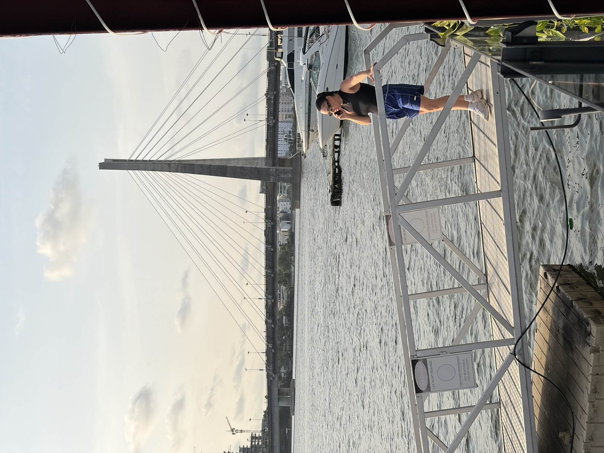 The image captures a daytime scene with a man standing on a white metal structure, possibly a dock or a platform, over a body of water. The man has a dark tank top and blue shorts and appears to be looking and possibly speaking into a phone. Behind the man and the platform, several white boats are seen docked on the water. In the background, there is a cable-stayed bridge.
In the very background, there are buildings and structures, potentially part of a cityscape. A sign is visible on the metal structure with the words "LIFE JACKET REQUIRED."
Based on the presence of the Ikoyi-Lekki Link Bridge, it appears that this photo was taken in Ikoyi, Lagos, Nigeria.