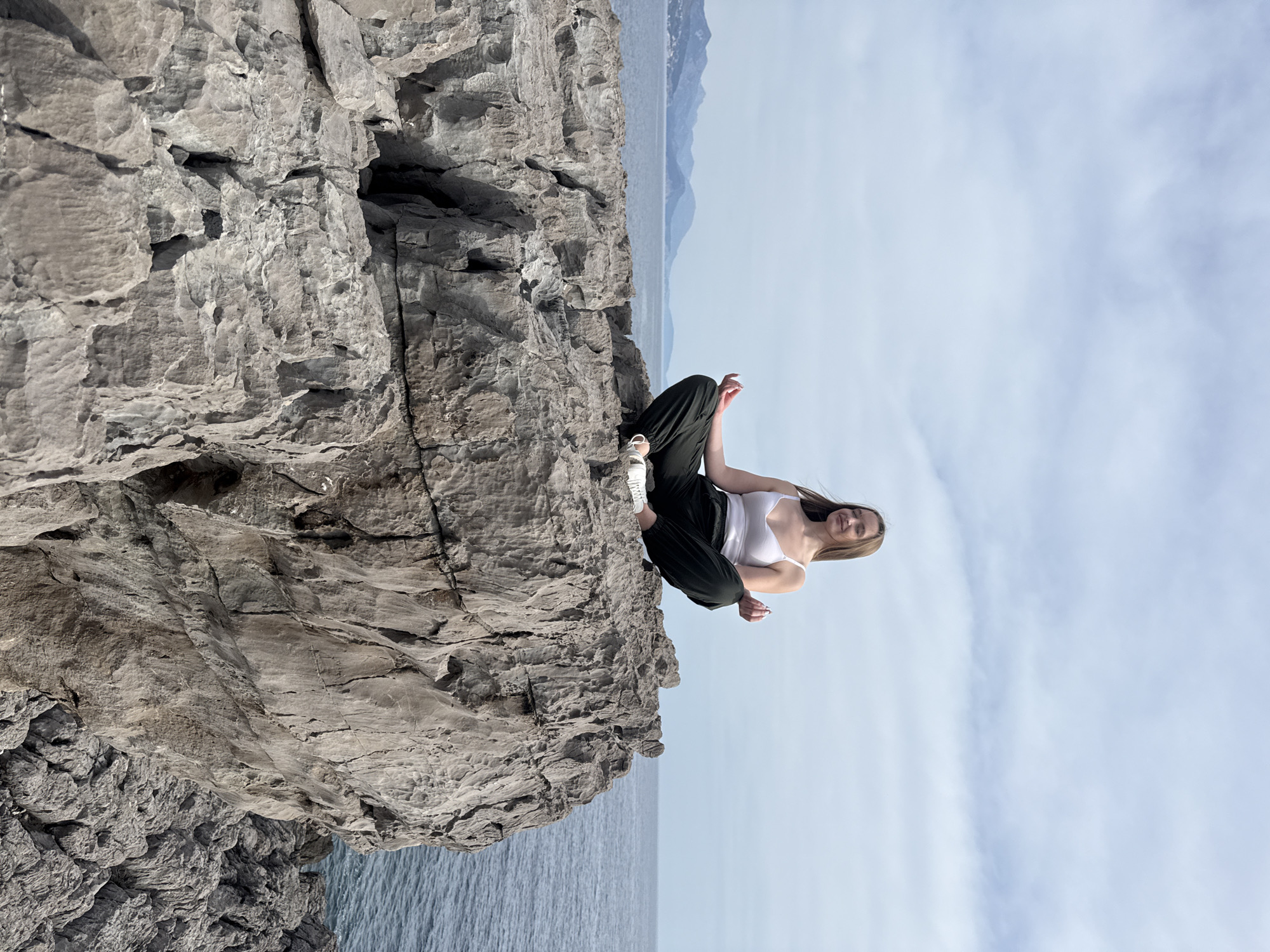 A young woman with long brown hair sits cross-legged on a rocky cliff in Boreti, Montenegro. She wears a white tank top and dark pants, with white sneakers visible near the edge of the cliff. The cliff is light gray and textured, with a small cave or opening visible on its surface. Below the cliff is a body of water, possibly the Adriatic Sea. In the distance, mountains are visible against a light blue sky with wispy clouds.