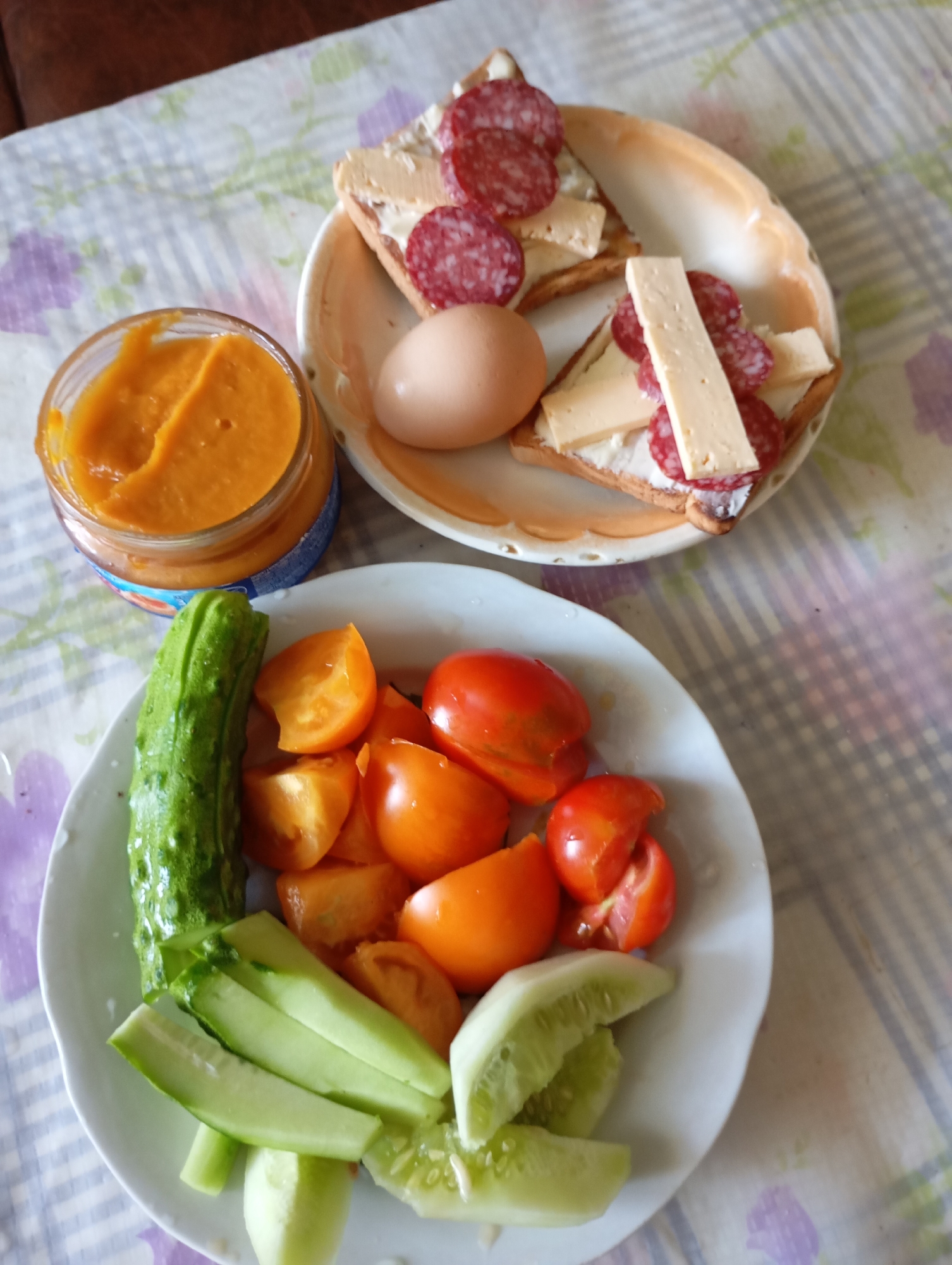 A close-up, top-down view shows a meal laid out on a checkered tablecloth. In the foreground, a white plate holds an assortment of chopped vegetables: a whole cucumber, sliced cucumber pieces, and chopped red and orange tomatoes. Behind this plate, to the left, is a small glass jar filled with an orange-colored spread, possibly pumpkin or carrot puree. To the right of the jar, on another slightly raised, scalloped-edged plate, are two open-faced sandwiches made with square crackers. Each sandwich is topped with a white spread, several thin slices of salami, and a strip of cheese. A single brown egg sits next to this plate. The setting appears to be a home environment, likely in Horishni Plavni, Ukraine, given the context. There are no people visible in the image. The lighting is natural and suggests an indoor setting.