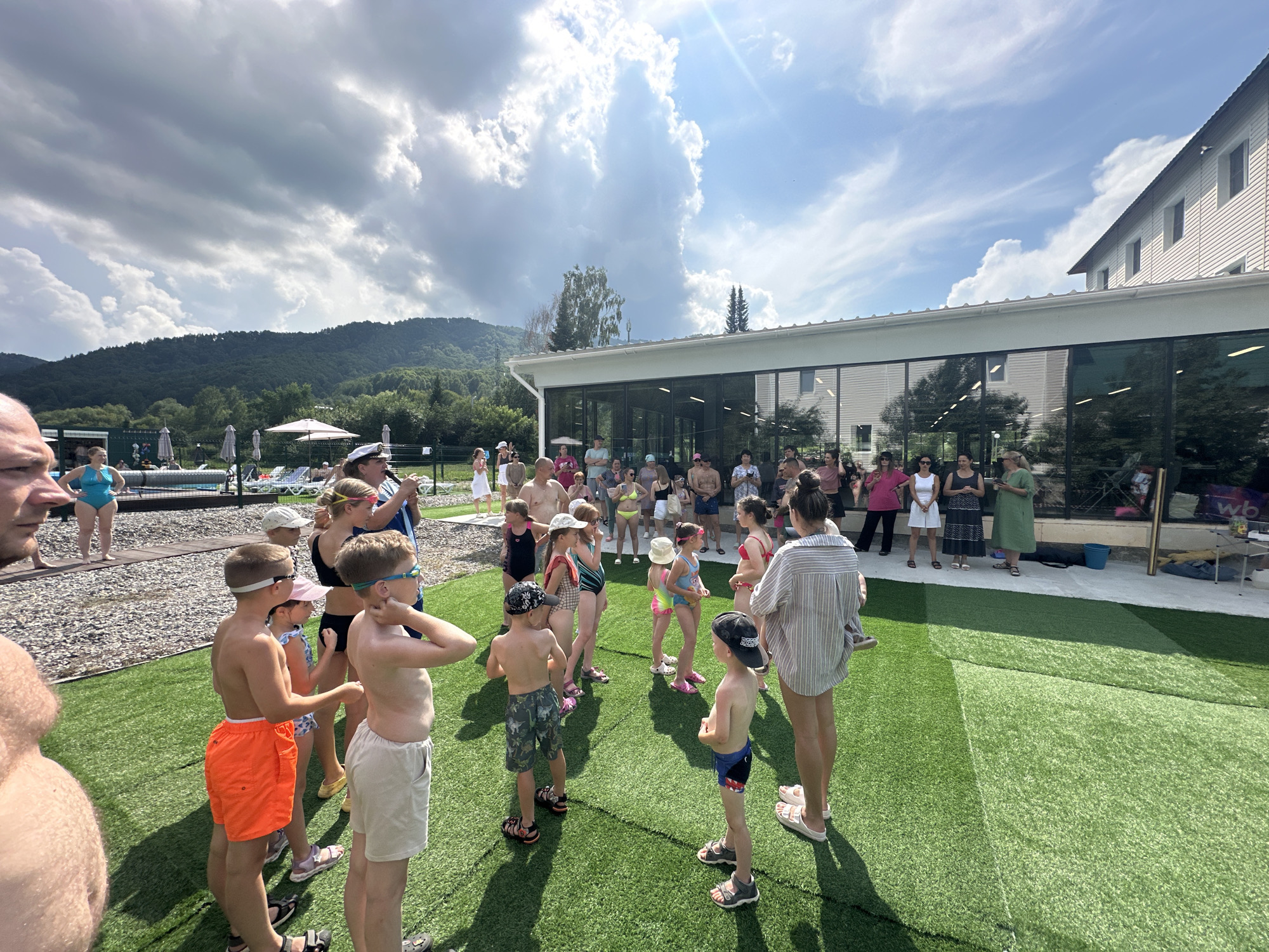 The image shows a group of people, primarily children and adults, gathered on a bright green artificial turf area next to a swimming pool. The setting appears to be a recreational facility, possibly a hotel or resort, with a building featuring large glass windows in the background. The sky is blue with dramatic white clouds, and a sunbeam breaks through. In the distance, a tree-covered mountain range is visible.
In the foreground, several children in swimwear are standing, some facing away from the camera, others in profile. They are wearing swim trunks and bathing suits, and some have goggles on. One boy in orange swim trunks is prominent on the left, facing left in profile. To his right, a boy in light-colored shorts stands facing forward, looking to the right. Another boy in blue and red swim trunks is seen from the back on the far right.
Behind the children, adults are also present. Some are watching the children, while others appear to be conversing. One woman in a striped shirt is standing on the far right, facing away from the camera, holding what looks like a small hula hoop.
The environment includes the artificial turf, a gravel area near the pool, lounge chairs, and umbrellas set up near the pool. The building in the background has a modern design with white siding and a prominent white roofline. The overall atmosphere suggests a sunny day at a resort or aquatic center. The specific location in Даниловка (алтайский), Russia, is not explicitly visible in the image but is stated as the context.