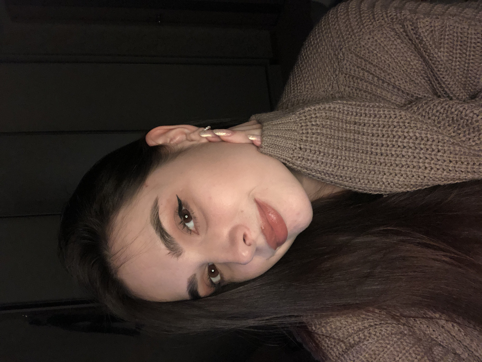 A young woman with dark hair, wearing a brown sweater, is lying on a dark surface. She is looking at the camera with her head tilted. She has fair skin, defined eyebrows, eyeliner, and her tongue is slightly out. Her left hand is near her ear, showing a ring and long, glittery nails. The background appears to be a dark, possibly indoor, setting. The location in the city of Noviy, Russia is unknown.