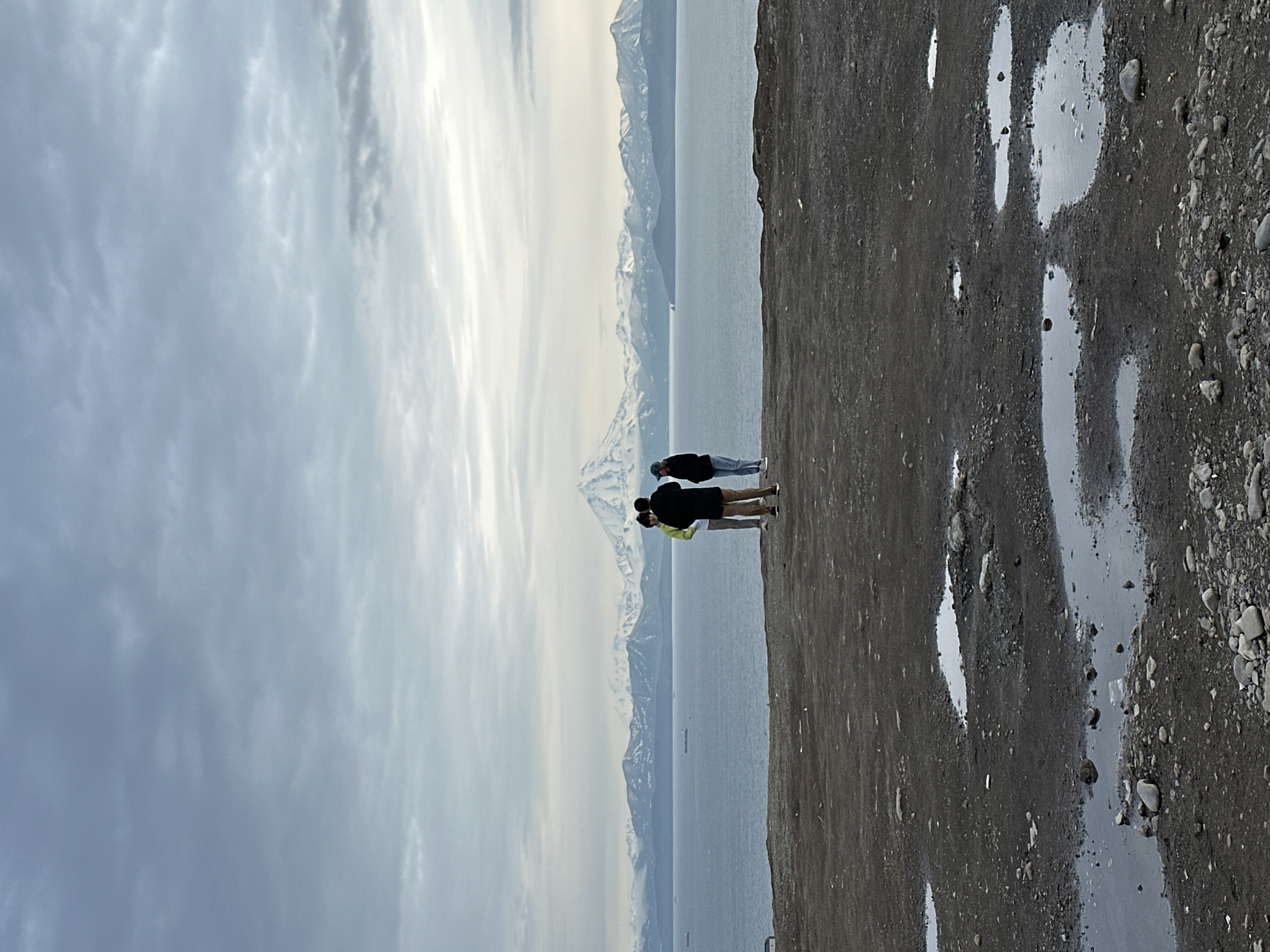Three individuals stand on a muddy, rocky overlook facing a large body of water. In the distance, a mountain range with snow-capped peaks is visible across the water. The sky is overcast with gray clouds. The foreground consists of dark, wet ground with scattered pebbles and several puddles. The overall scene suggests a natural, rugged landscape under a cloudy sky. The image is likely taken from a vantage point overlooking the Avacha Bay in Petropavlovsk-Kamchatsky, Russia, given the presence of snow-capped volcanoes characteristic of the region.