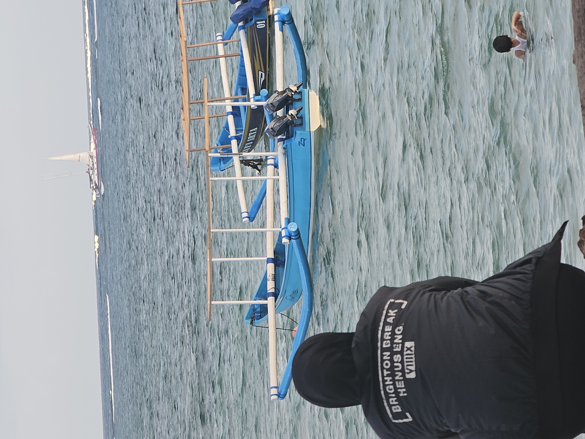 A person wearing a black hooded jacket with white lettering on the back, reading "BRIGHTON BREAK HENUS ENG. VIIIMIX", is sitting on a stone ledge, facing the ocean. The jacket has a prominent hood obscuring the person's head. In the foreground, the water is choppy and a dark blue. In the mid-ground, two blue and white outrigger boats are moored in the water. The boat closer to the viewer has two outboard motors attached to its stern. Further out in the ocean, a large sailboat with white sails is visible, along with a few smaller boats or buoys. The sky is overcast and pale. In the far right of the image, a young person wearing a white tank top is swimming in the ocean. This scene is consistent with a location along the coast of Denpasar, Indonesia, likely a beach or harbor area.