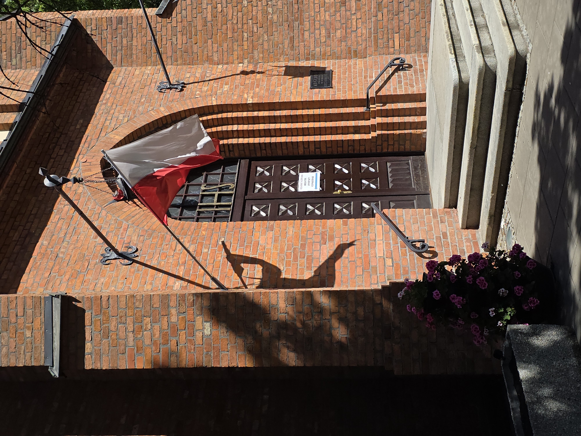 The image shows a brick building in Warsaw, Poland, with a Polish flag hanging to the left of a dark wooden door. Above the door are brick steps leading upward, with metal handrails on either side. A plaque is visible on the wall above the steps. To the right of the doorway are light-colored vertical architectural elements and a planter filled with pink flowers. Shadows are cast across the scene, including a distinct shadow of a person standing near the photographer.