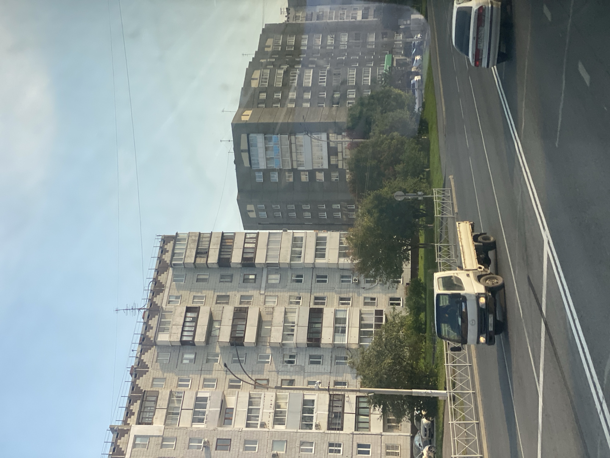 The image depicts a street scene in Kemerovo, Russia, likely during daytime with clear skies. Two large, multi-story residential buildings dominate the background. The building on the left is light gray with numerous windows and balconies, featuring a decorative patterned top. A tall street lamp is visible in front of it. The building on the right is darker gray, also with many windows and balconies.
In the foreground, a white flatbed truck with a black cab is driving on a multi-lane road. A white sedan is also visible on the road, further ahead and to the right. The road is marked with white lane lines. A metal fence separates the road from a grassy area with trees behind it. Several cars are parked on the side of the road in the distance, near the buildings. The overall impression is a typical urban residential area.