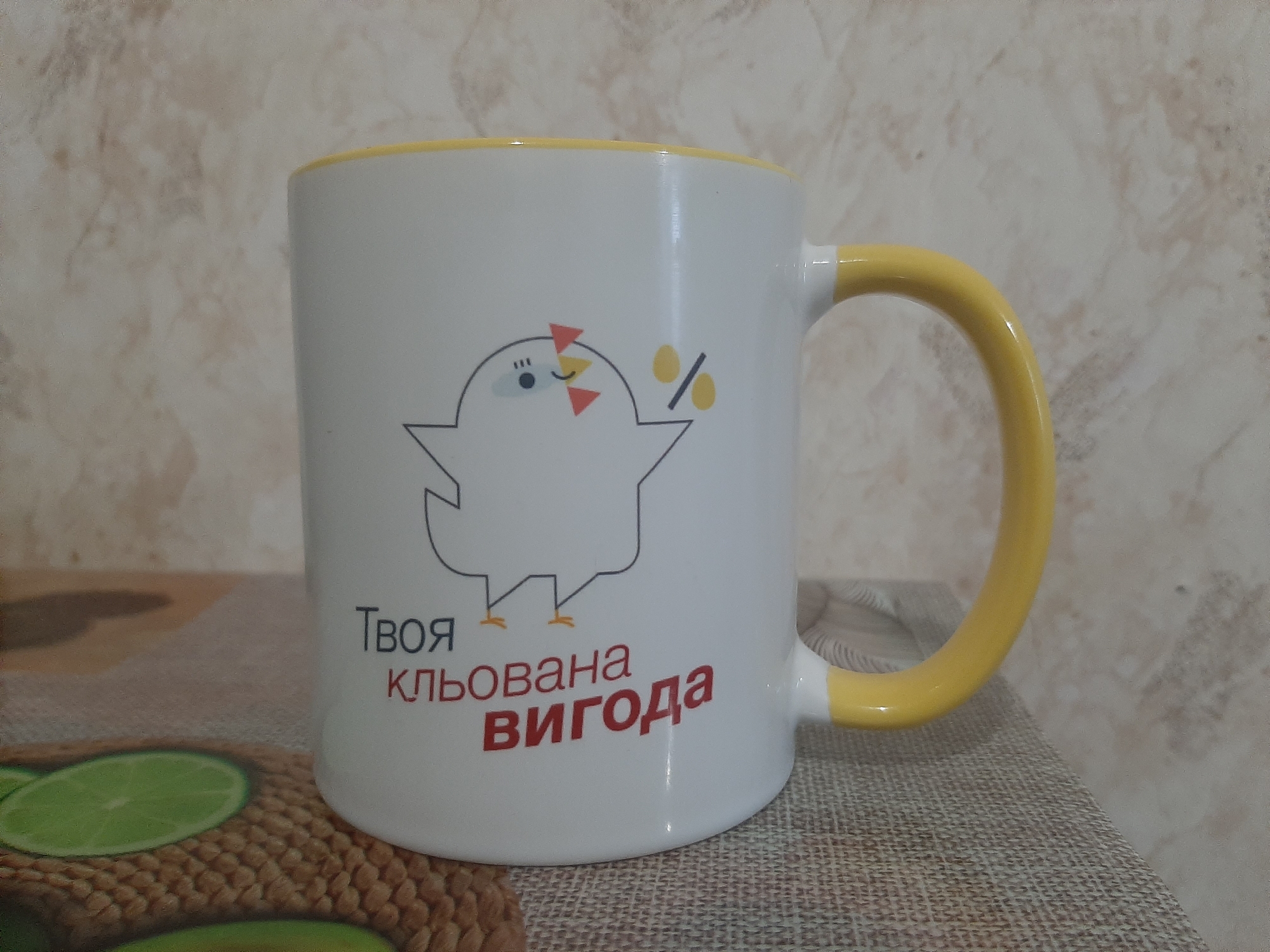 A white ceramic mug with a yellow handle and rim is shown in a close-up, centered shot. The mug features a cartoon drawing of a stylized bird or chick with a beak and closed eyes. Above the bird's head are small orange triangles and yellow circles with percentage symbols. Below the bird, text is printed in two lines: "Твоя" (Your) in dark grey and "КЛЬОВАНА ВИГОДА" (Beaked Benefit) in red. The mug rests on a textured surface, possibly a placemat, with a few green lime slices visible on the left side of the frame. The background is a textured wall with a light beige and brown pattern. The scene is well-lit and appears to be an indoor setting.