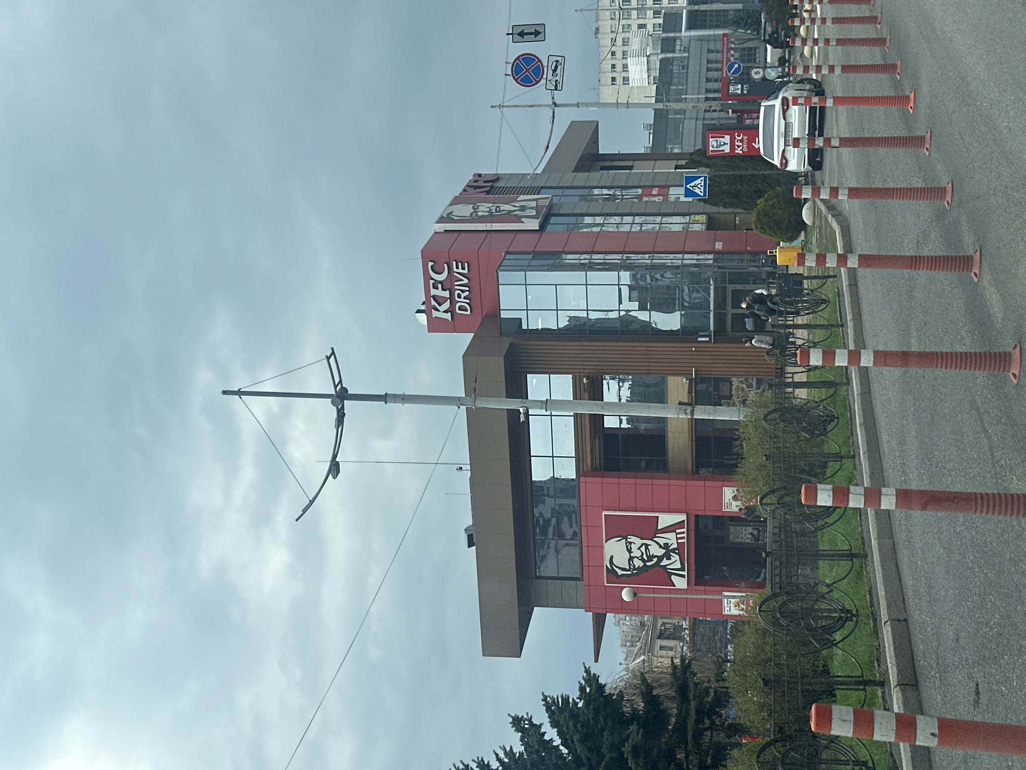 A street-level view shows a KFC Drive restaurant in Kyiv, Ukraine. The restaurant is a multi-story building with a red and glass facade. The KFC logo is prominently displayed. A row of orange and white striped traffic cones lines the street. Trees and a low fence are visible in front of the restaurant. Street signs are posted on poles near the building. A light gray sky is overhead.