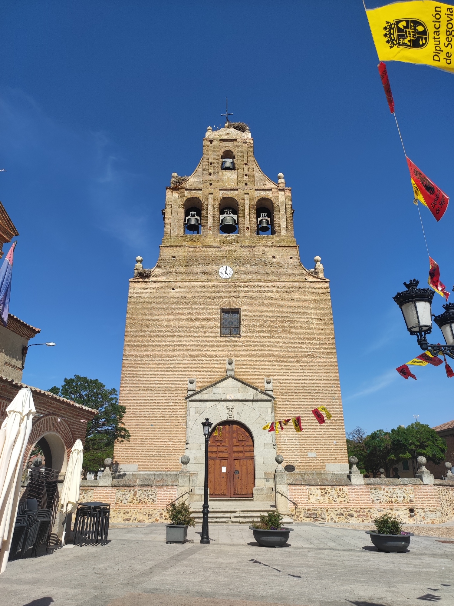 The image shows the bell tower of a brick church in Sangarcía, Spain. The tower has arched openings with bells and a clock face. Flags are flying from the top of the tower and from lampposts in the foreground. There are also some potted plants and stacked chairs in the scene. The sky is clear and blue.