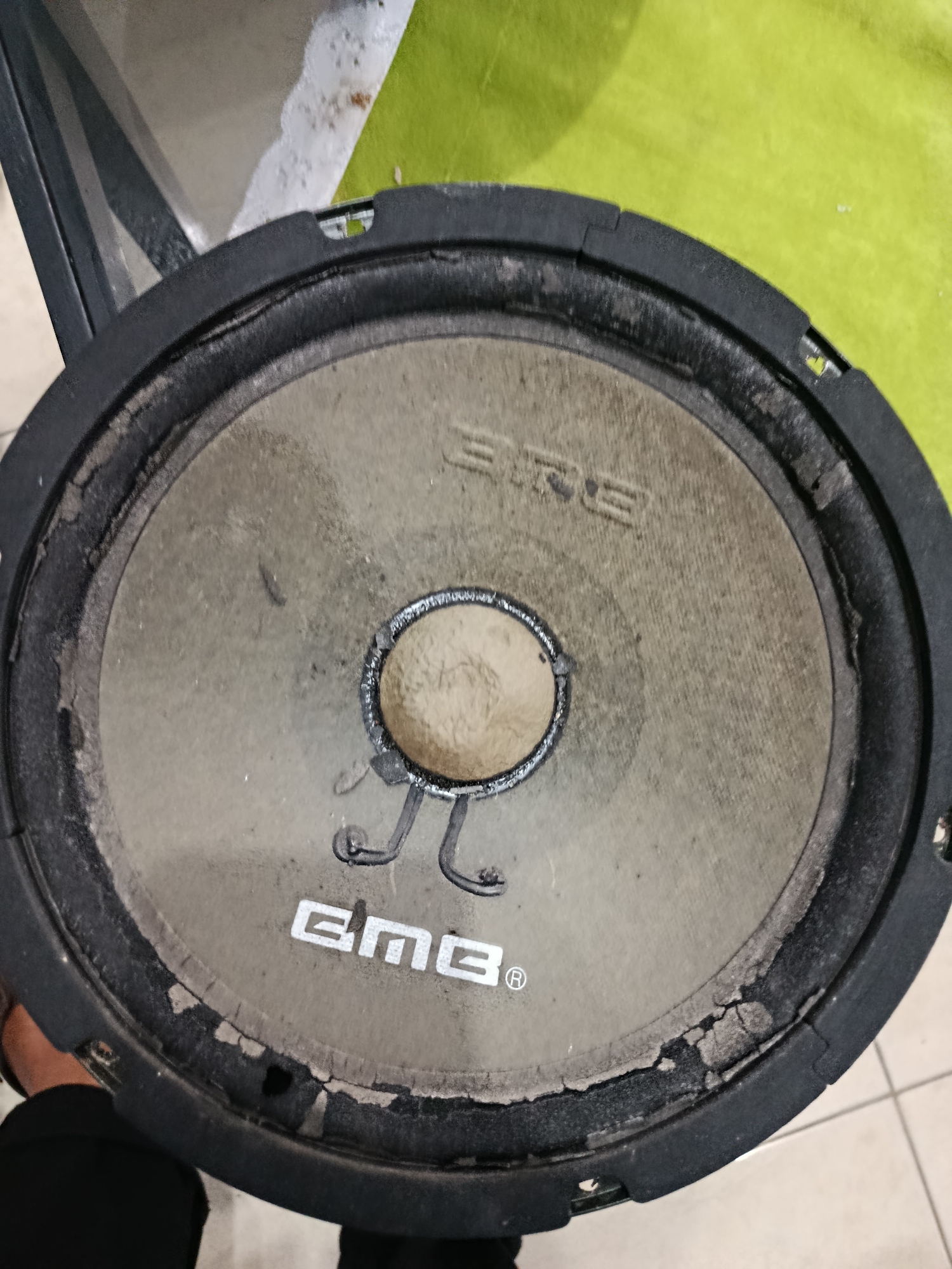 A close-up, overhead shot shows a damaged speaker. The speaker has a textured, light-colored cone with a circular, textured dust cap in the center. The words "EMG" are visible in white, stylized lettering on the cone. The outer edge of the speaker, which would typically be a flexible surround, is severely degraded, with pieces of material flaking and missing, exposing the inner workings. The speaker is framed by a black, rigid basket. In the background, a green surface and a tiled floor are visible. No specific location in Palembang, Indonesia can be determined from the image.