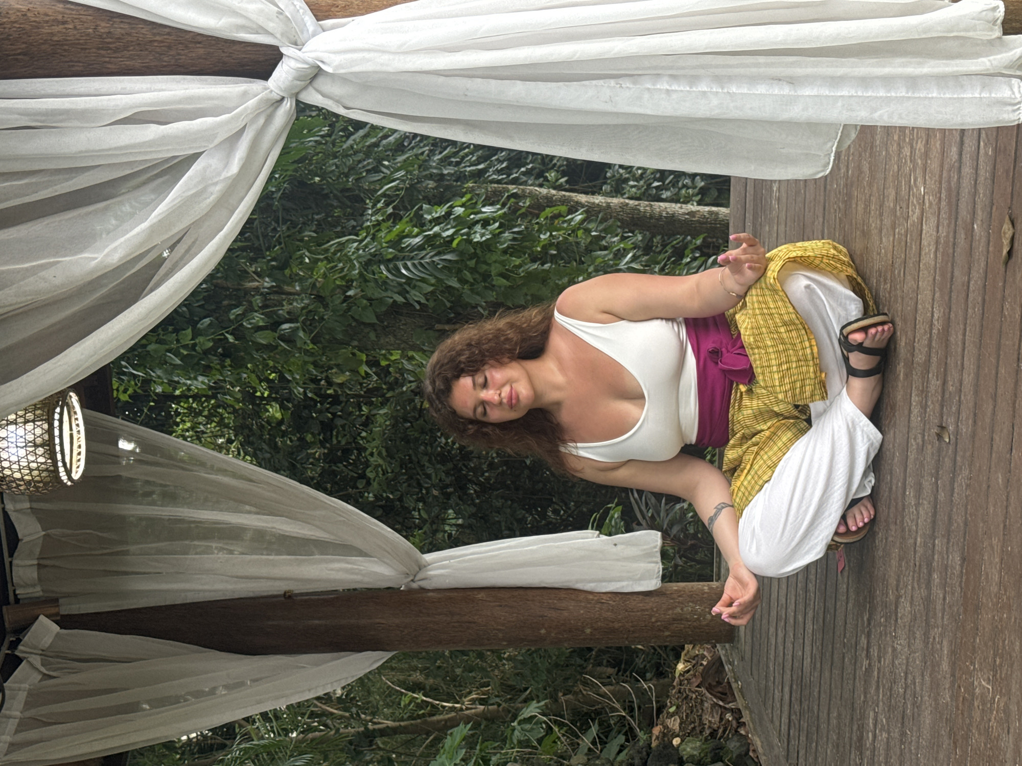 An outdoor, vertical-oriented photograph captures a woman meditating or relaxing on a wooden deck amidst lush tropical greenery in the Ubud District, Indonesia. The woman, with curly brown hair, wears a white tank top, a magenta wrap-around skirt, and white wide-leg pants, complemented by dark sandals. Her hands are gently posed, possibly in a mudra, and her eyes are closed, conveying a sense of peace and calm. She has light pink nail polish on her fingers and toes, and a tattoo is visible on her right forearm.
The scene is framed by white, sheer fabric draped and tied around rustic wooden beams, creating an ethereal canopy effect. A woven light fixture or lantern with a warm glow is visible on the left side of the frame, suggesting the time might be late afternoon or early evening. The background is dominated by dense, verdant foliage and trees, indicating a natural and secluded setting. The wooden deck beneath her is made of vertical planks, adding to the natural aesthetic. No visible text is present in the image.