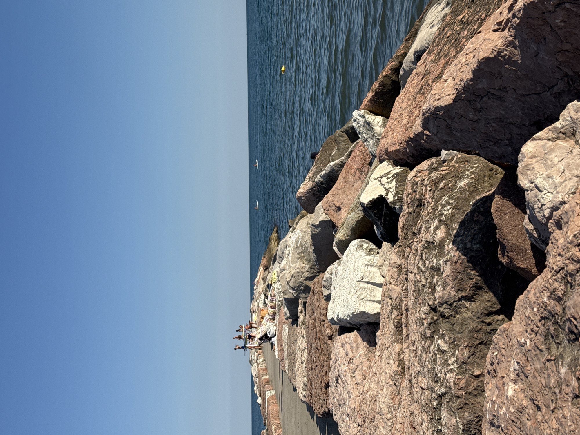 The image depicts a breakwater made of large, rough, reddish-brown and white rocks leading into a calm, deep blue sea under a clear, light blue sky. On the breakwater, in the distance, a small group of people are visible. Some are standing and appear to be interacting, while others are seated or lying down on towels. Further out on the water, there are a few individuals on paddleboards, and a yellow buoy is floating. The overall scene suggests a sunny, recreational day at a coastal location in Eraclea, Italy.