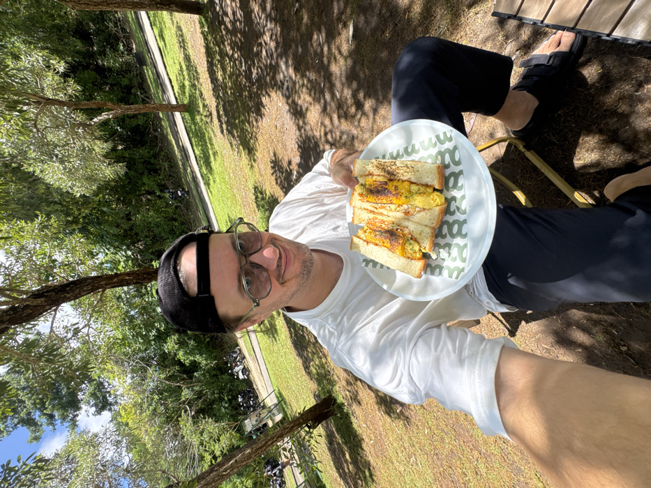 A person wearing a white long-sleeved shirt, dark pants, a black cap, and glasses holds a plate of sandwiches. The sandwiches are cut into rectangles with a filling visible between the bread slices. The plate has a green and white geometric pattern. The person is seated outdoors, with trees and a grassy area visible in the background. The lighting suggests a sunny day, with shadows cast on the ground.