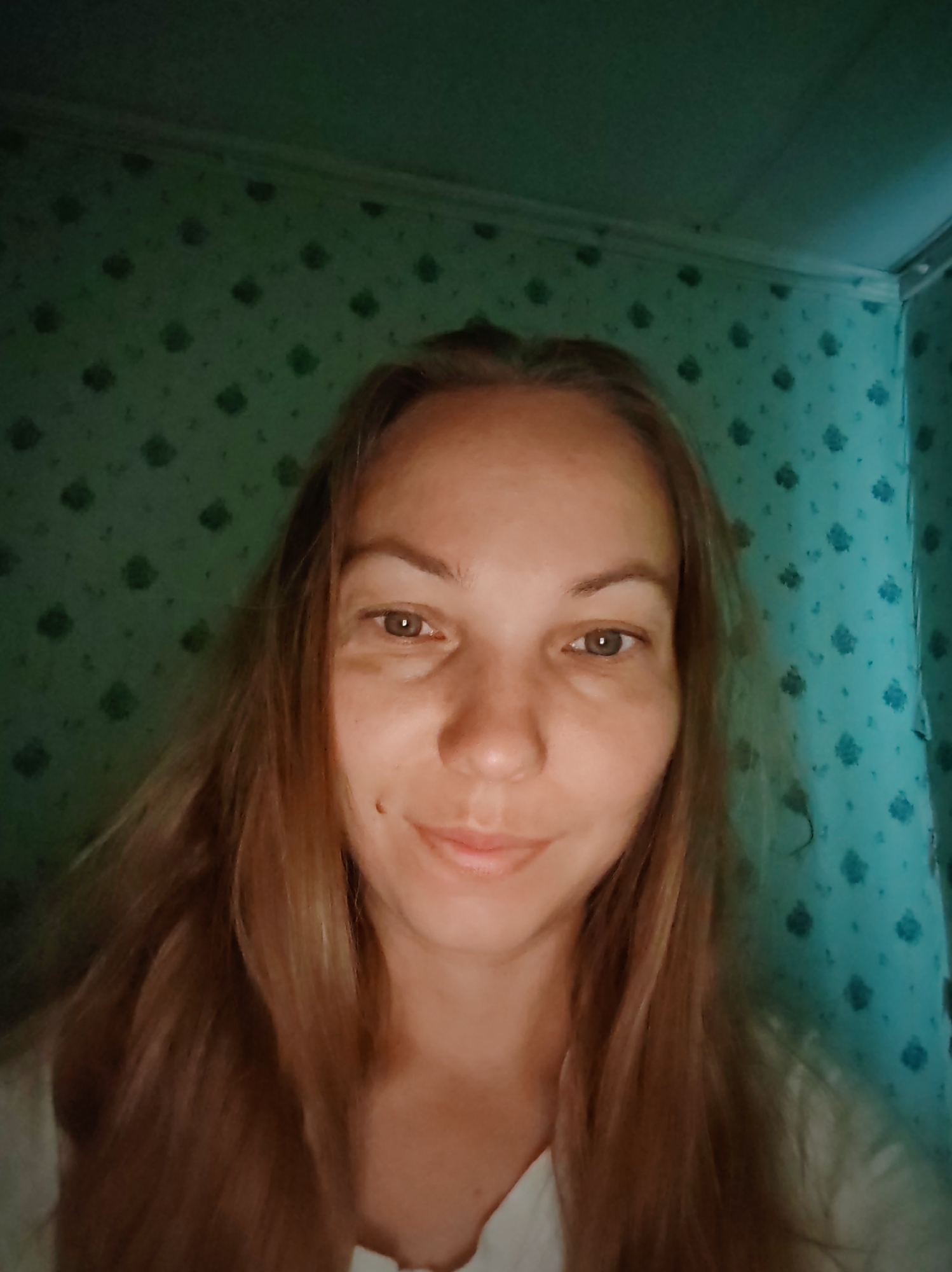 The image shows a medium shot of a woman with long, wavy, light brown hair. She has light brown eyes and is looking directly at the camera. She is wearing a white shirt. The background appears to be a room with patterned green wallpaper and a white trim at the top. The lighting suggests an indoor setting. The image was taken in Chukhlem, Russia.